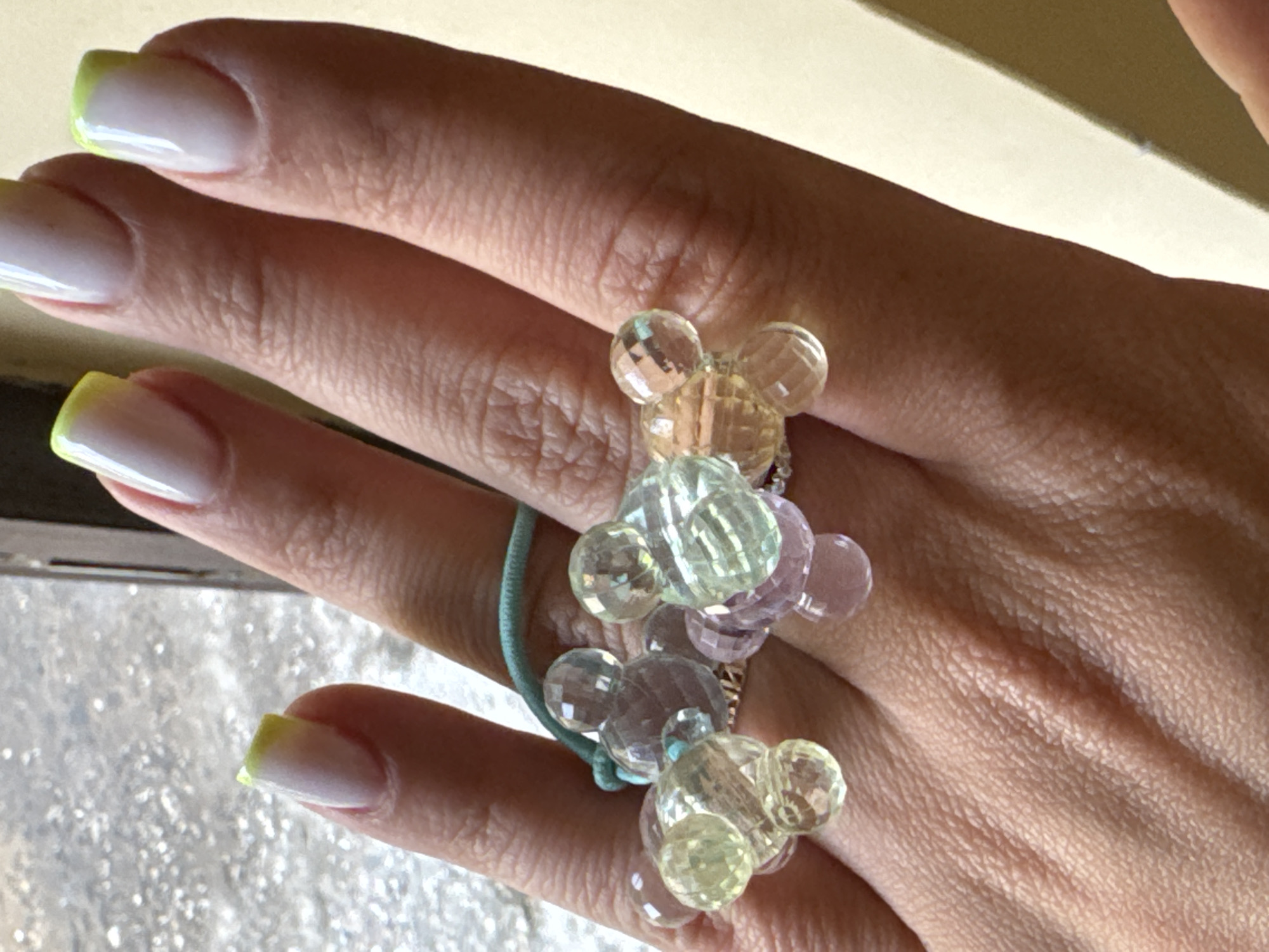 A close-up shot focuses on a person's hand adorned with a multi-colored, Mickey Mouse-shaped beaded ring. The ring features an assortment of faceted beads in pastel shades of pink, yellow, green, and clear, forming clusters of the iconic character's head and ears. The hand has well-maintained fingernails with a French manicure, featuring a white base and a vibrant yellow tip. The skin tone suggests the image was taken in natural lighting, possibly indoors given the soft, diffused illumination and lack of strong shadows. The background is out of focus, with a pale, yellowish surface at the top and a blurred, textured area at the bottom, suggesting a casual indoor setting. There is no visible text in the image. The overall impression is one of feminine adornment and a playful, whimsical aesthetic.