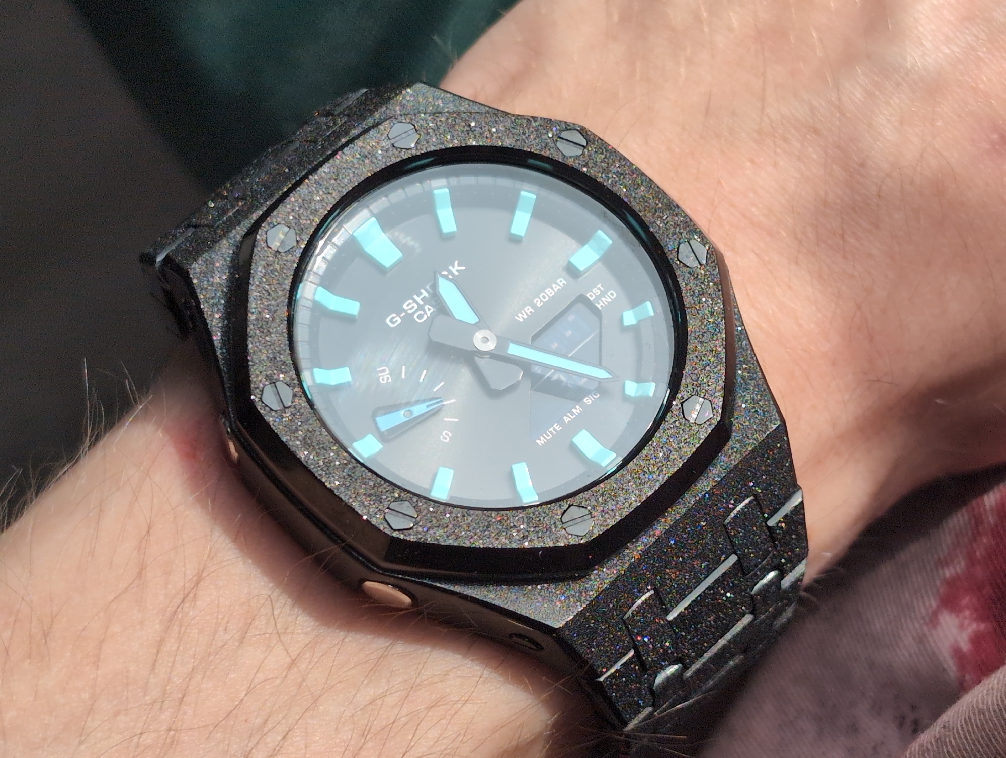 A wrist wearing a G-Shock watch is shown in close-up. The watch has a dark gray, octagonal case with a glittery finish and eight screws around the bezel. The watch face is dark gray with light blue hour markers and hands. The band is also dark gray with a glittery finish and a brick-like pattern. The wrist appears to be fair-skinned with light hair. There is no indication the image was taken in Izhevsk, Russia.