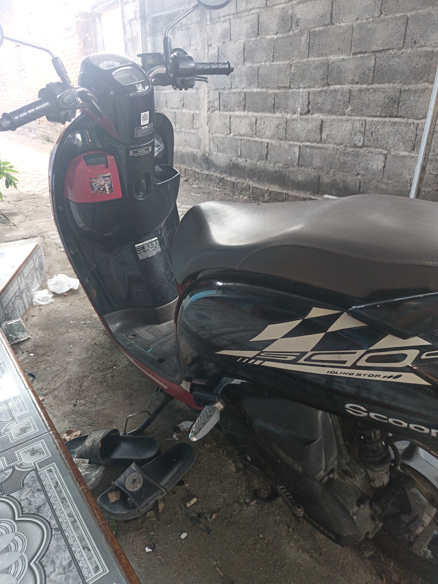 A red and black Honda Scoopy motorcycle is parked outdoors, possibly in a yard or alleyway in Temuwulan, Indonesia. The motorcycle is positioned at an angle, with the front facing left and slightly towards the viewer. A brick wall, weathered and discolored, forms the backdrop behind the motorcycle. In the foreground, a black patterned table or counter edge is visible to the left. A pair of black slide sandals are placed on the ground near the motorcycle's rear wheel. The ground surface appears to be dirt or gravel, with some scattered debris including white plastic and cigarette butts. The lighting suggests it is daytime, with bright sunlight casting shadows and creating some glare on the motorcycle's bodywork. The scene is static, with no people or immediate action depicted. The overall impression is of a parked vehicle in a somewhat unkempt outdoor setting.