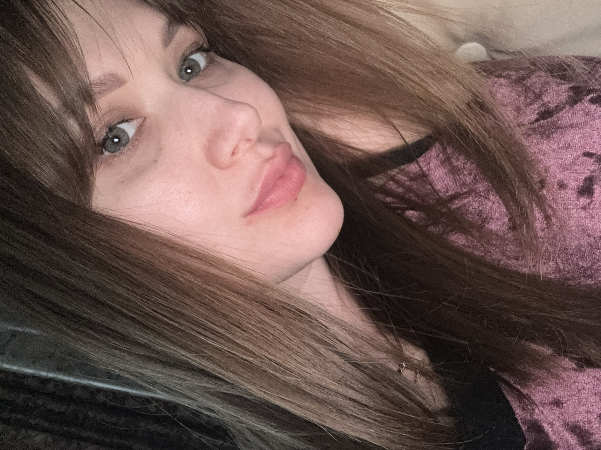 A close-up, angled portrait of a woman with fair skin, light gray-green eyes, and brown hair with lighter highlights. She has bangs that partially cover her forehead. Her lips are full and appear to have a slight pink tint. She is wearing a textured, pinkish-purple top with a black undershirt visible at the neckline. A thin, gold-colored necklace with a small pendant is partially visible. The background is dark and out of focus. There is no information indicating the location is Chaikovsky, Russia.