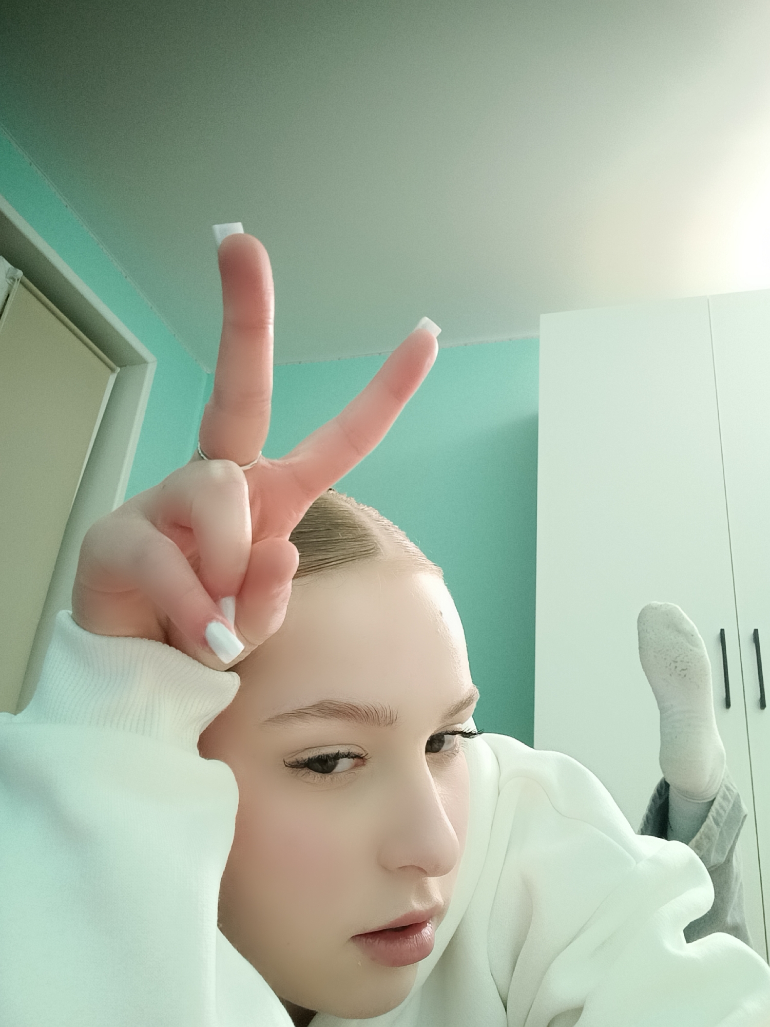 A person with light skin and light brown hair is reclining, wearing a white sweatshirt and white socks. Their left hand is raised, making a peace sign. The background includes a light green wall and a white wardrobe. The location is a room interior, likely within the city of Chaikovsky, Russia.