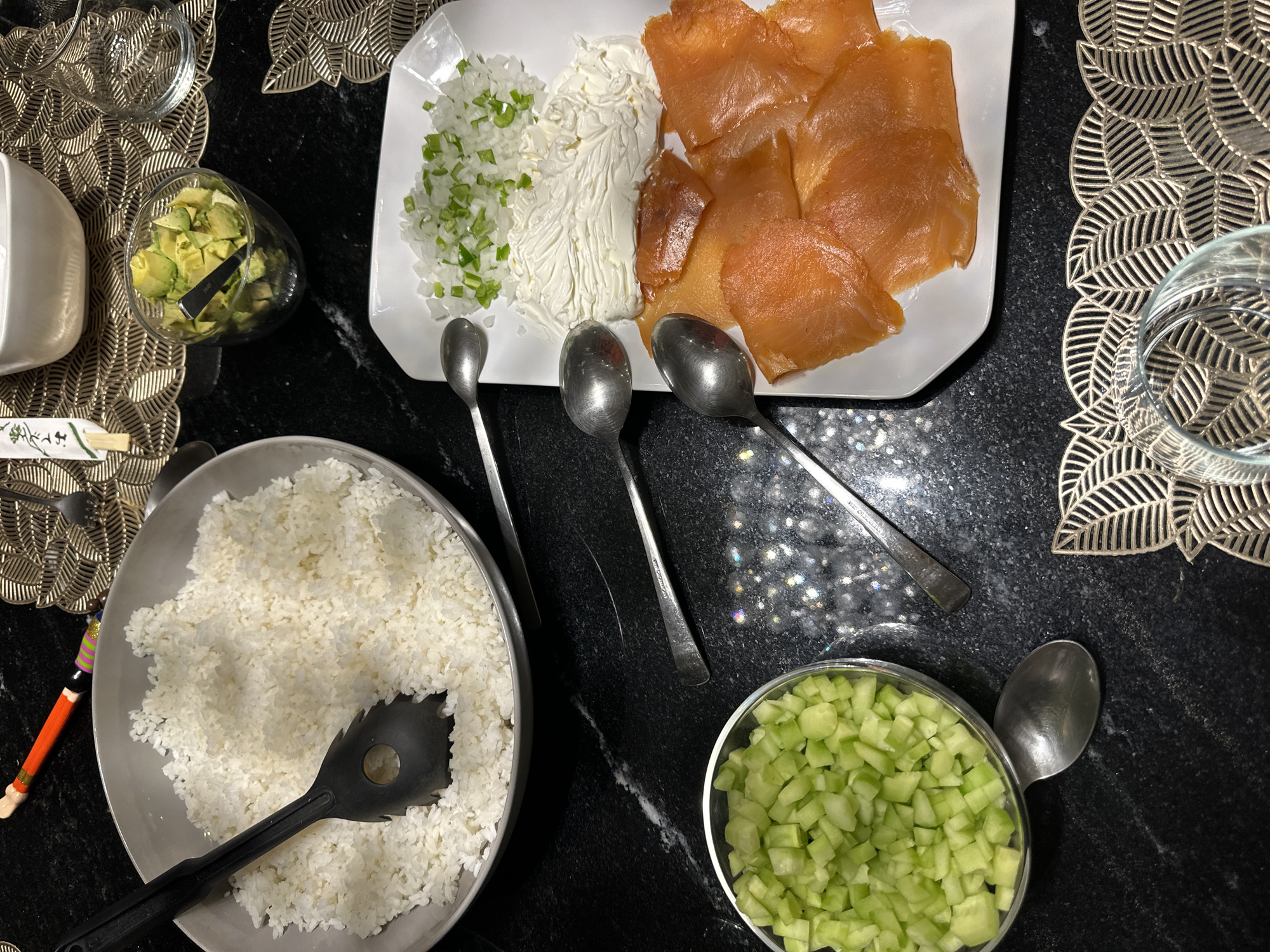 A table setting with various food items is presented against a black, speckled surface resembling granite. A large bowl of cooked white rice sits on the left side of the frame with a black serving utensil inside. Adjacent to it, a glass bowl contains diced green cucumbers, with a spoon resting beside the bowl.
On the top right of the table, a white rectangular plate holds three separate components: diced onions and green peppers, a pile of white cream cheese, and a stack of sliced salmon. Three spoons are arranged below the plate.
To the upper left, a glass jar is filled with diced avocados, with a spoon inside. A pair of chopsticks rests near the avocado jar. A clear glass is positioned on the right side of the frame, and decorative gold leaf-shaped objects are present on both sides.
The setting appears to be indoors, and no people are visible in the frame. There is no other indication about the location in Mexico City, Mexico.