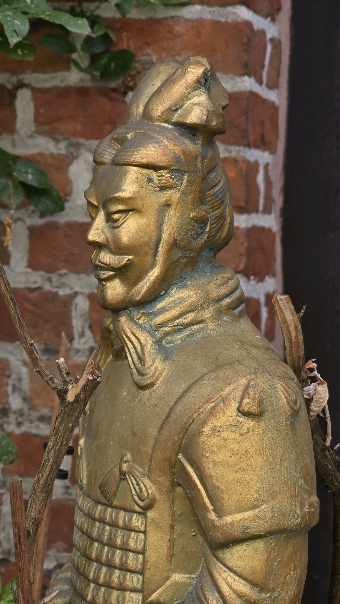 The image shows a close-up of a golden statue of a Terracotta Army warrior. The statue is partially obscured by a thin, dry branch in the foreground and some greenery on the left. The warrior is depicted from the chest up, with detailed armor, a mustache, and a stern expression. The background consists of a brick wall and a dark, out-of-focus area. The statue appears to be an outdoor decoration, possibly in a garden or courtyard. The statue is located in Antwerp, Belgium, at the corner of Kipdorpbrug and Keizersbrug, next to the Red Star Line Museum, in the vicinity of a playground.