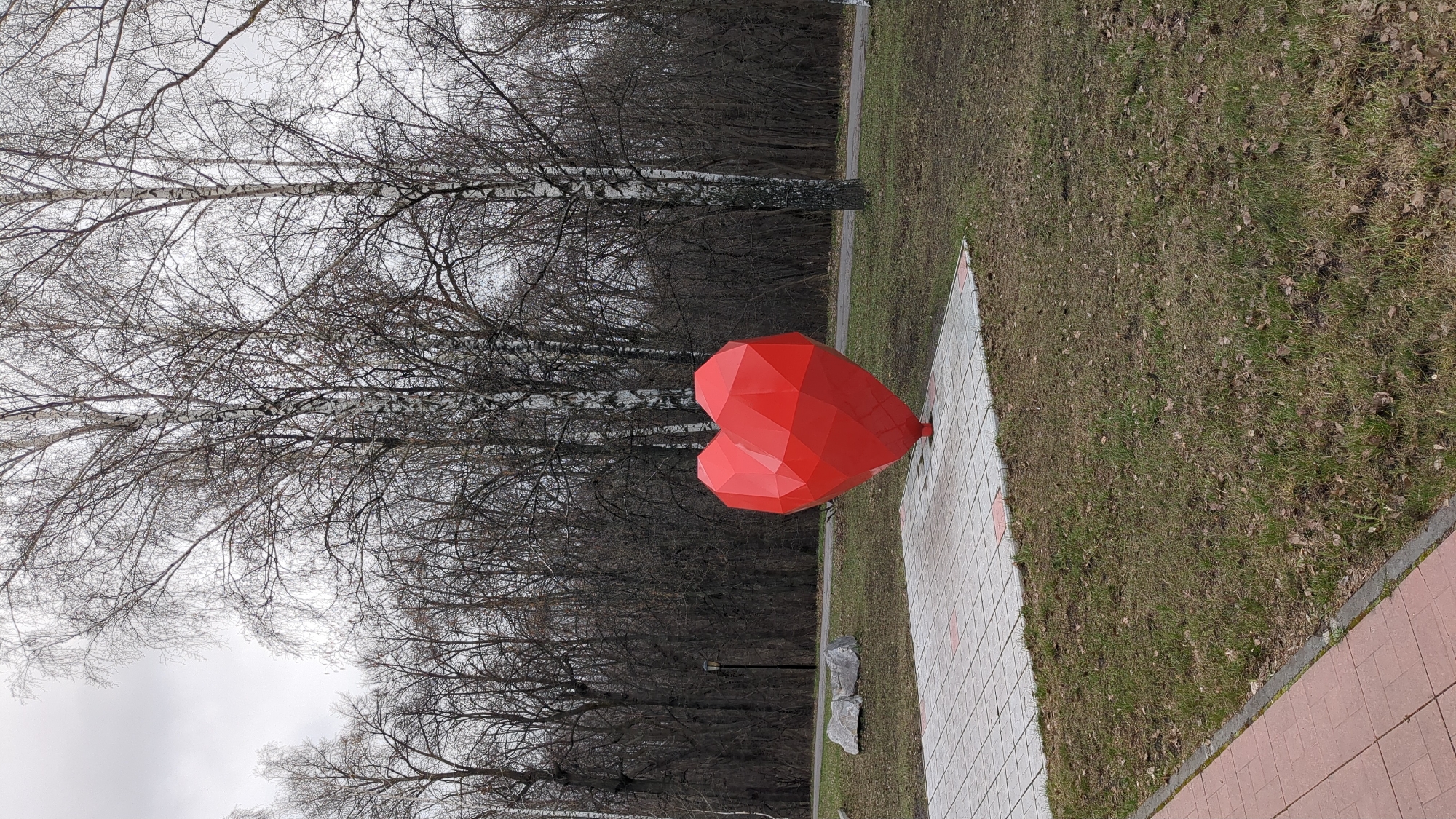 The image depicts a red, geometric heart sculpture situated on a tiled walkway next to a grassy area in a park. Behind the heart are bare trees, characteristic of late autumn or early spring. The trees form a dense backdrop, suggesting the edge of a forest or a heavily wooded area. The sky is overcast, creating a muted lighting condition. The location is indicated as Ulyanovsk, Russia. The scene is devoid of people or any visible actions or interactions.