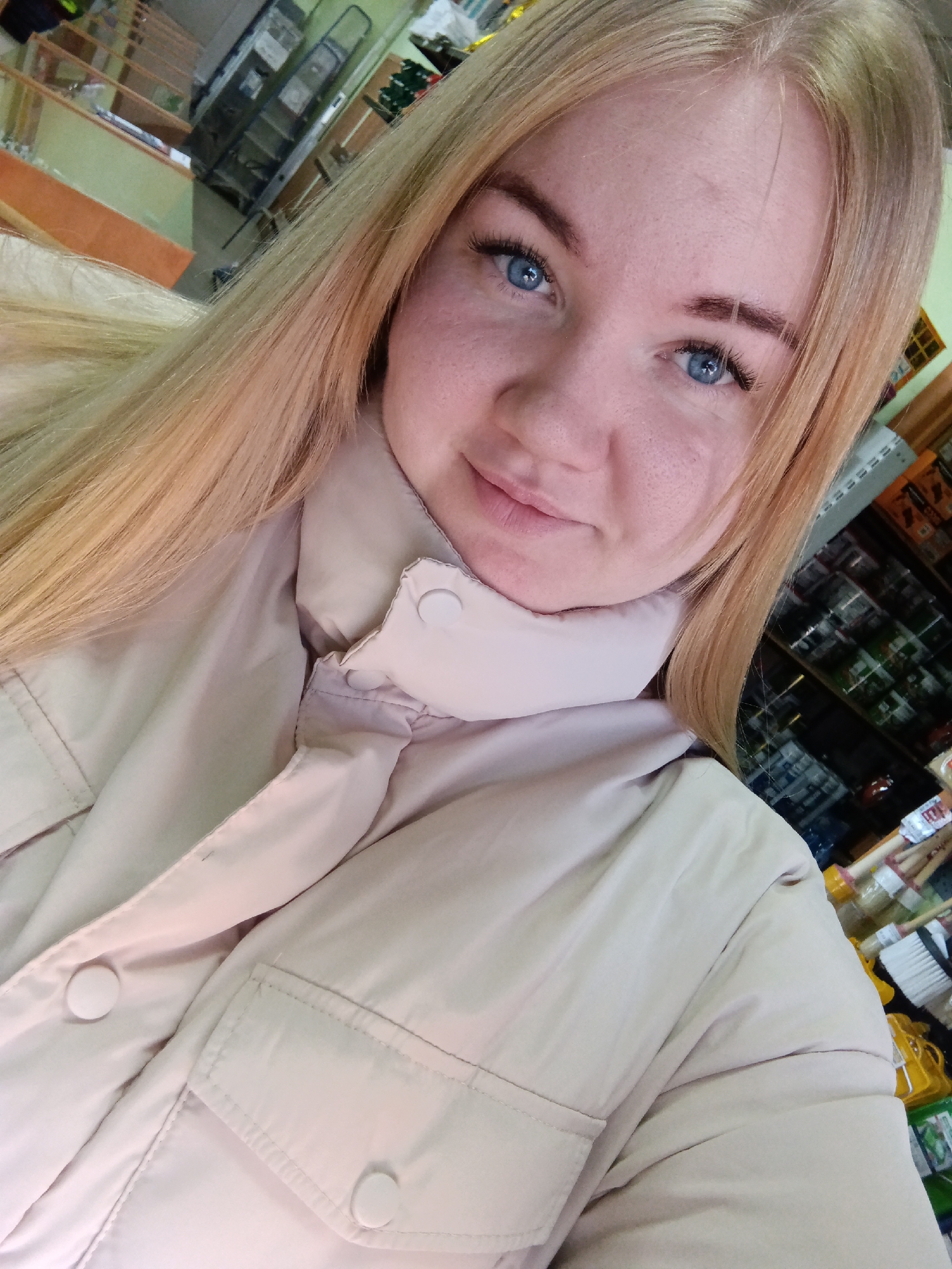 A woman with fair skin, blue eyes, and long blonde hair is looking at the camera. She is wearing a light beige jacket. Shelves filled with various items are visible in the background. The location appears to be a store. The city is not explicitly stated as Фоки in the image.