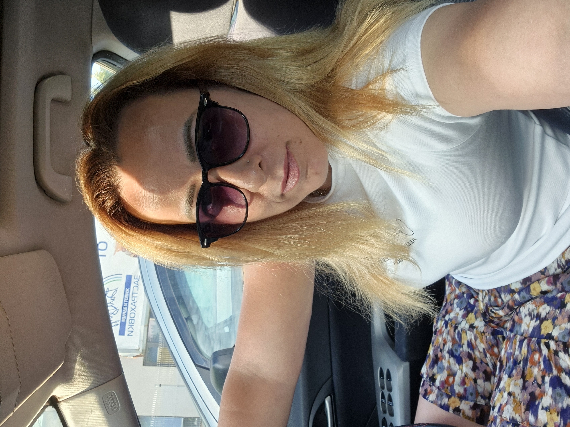 A woman with blonde hair and sunglasses is taking a selfie in the driver's seat of a car. She is wearing a white t-shirt and a floral skirt. The car's interior is visible, including the dashboard and a window. A sign with text is visible through the window. The overall lighting suggests it is daytime.