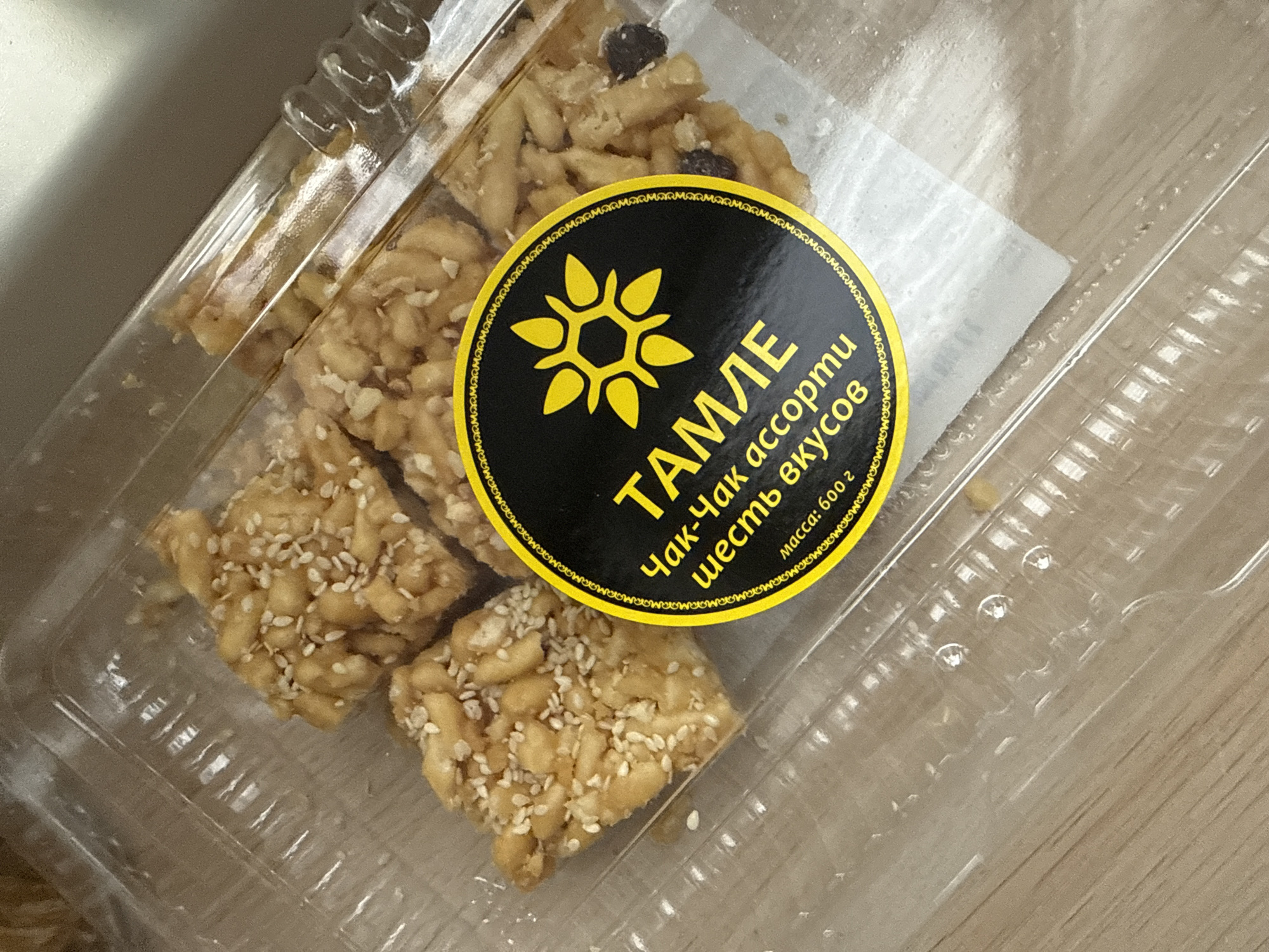 A close-up shot depicts a clear plastic container filled with what appears to be an assortment of fried dough pastries, commonly known as Chak-Chak. The pastries are coated with sesame seeds. A round label is affixed to the packaging. The label is black with a yellow border. On the label is the word "TAMLE" in large, yellow Cyrillic letters. Beneath this is the phrase "Чак-Чак ассорти шесть вкусов" which translates to "Chak-Chak assortment six flavors." There is also a stylized sunflower or starburst symbol in yellow on the label. Below is the text "масса: 600 г" which translates to "mass: 600 g." The scene appears to be indoors, with a light-colored wooden surface underneath the container. The specific location within Moscow, Russia, cannot be determined from the provided image.