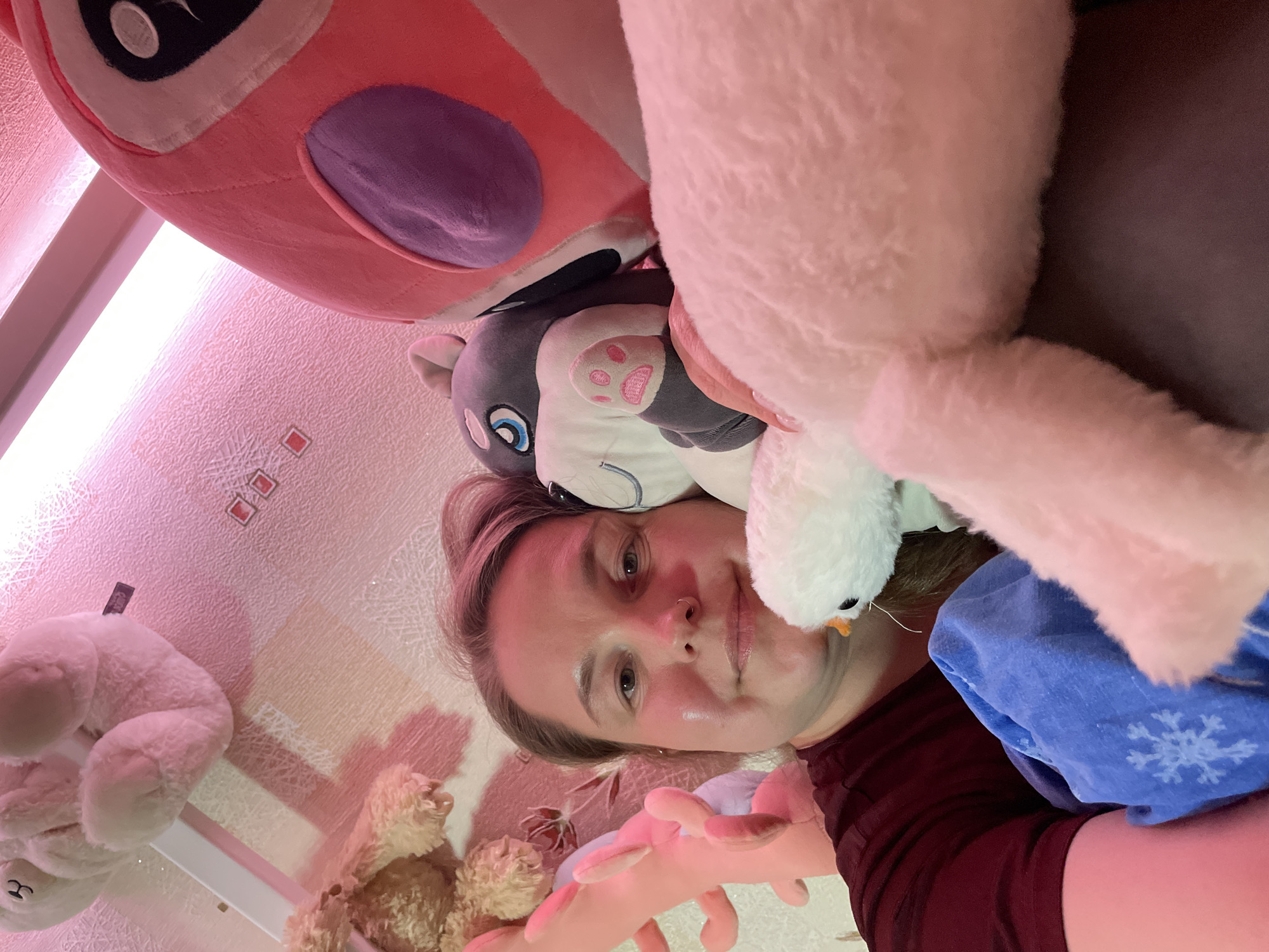 A person is lying down, face-up, surrounded by stuffed animals. One large pink and purple plush toy is near the top of the frame. Several other plush toys, including a gray and white husky-like toy with pink paw prints and a small white bird plush toy, are positioned around the person’s face and head. A blue piece of fabric with a snowflake design is also present. In the background, a patterned wall is visible, along with additional stuffed animals, including teddy bears, on a shelf. The location is Izhevsk, Russia.
