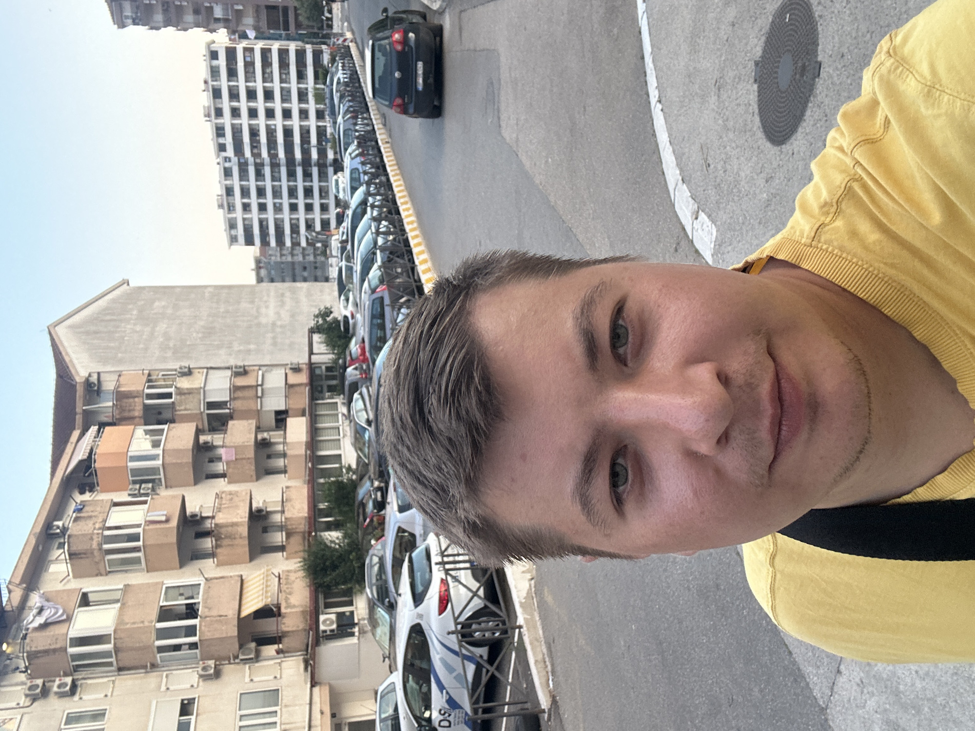 This is a portrait-style selfie taken outdoors with a shallow depth of field, focusing on the face of a young man wearing a yellow t-shirt. The man has light skin, short brown hair, and is looking towards the camera with a neutral expression. His right eye is clearly visible and is green.
The background is blurred but suggests an urban setting. To the left, there are apartment buildings with visible balconies and air conditioning units, characteristic of Mediterranean architecture. To the right, a row of parked cars lines a street, with some vehicles appearing to be police cars, indicated by blue and white markings. A concrete sidewalk and a street are also visible, with a circular manhole cover on the pavement.
The lighting suggests it is daytime, likely morning or afternoon, with bright sunlight. The weather appears to be clear and warm. The presence of parked cars and apartment buildings indicates a residential or commercial area within a city, specifically Budva, Montenegro. The selfie perspective, with the person's face taking up a significant portion of the frame and the background out of focus, is typical of this photographic style. No legible text is clearly visible in the image.