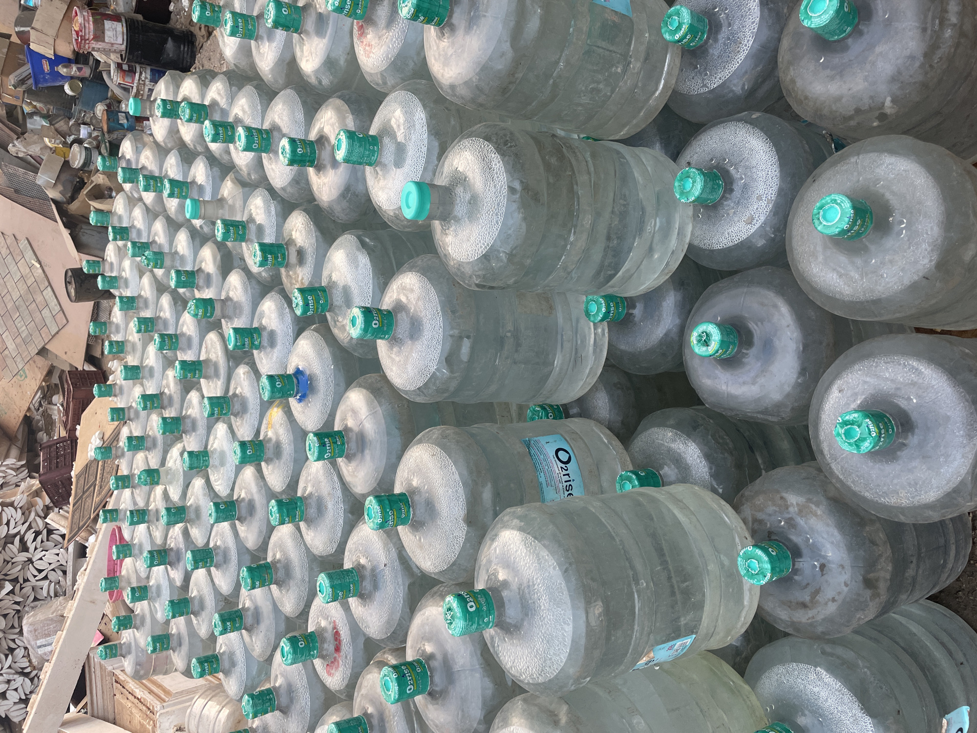 A tightly packed stack of numerous large, transparent water bottles, each with a distinctive green cap bearing the label "O2rise," dominates the frame. The bottles are arranged in a grid-like pattern, creating a dense wall of plastic containers. To the left of the bottles, a collection of assorted debris is visible, including discarded materials and remnants of construction or demolition, with paint cans. The overall setting suggests an outdoor storage area in what appears to be Mira-Bhayander, India.