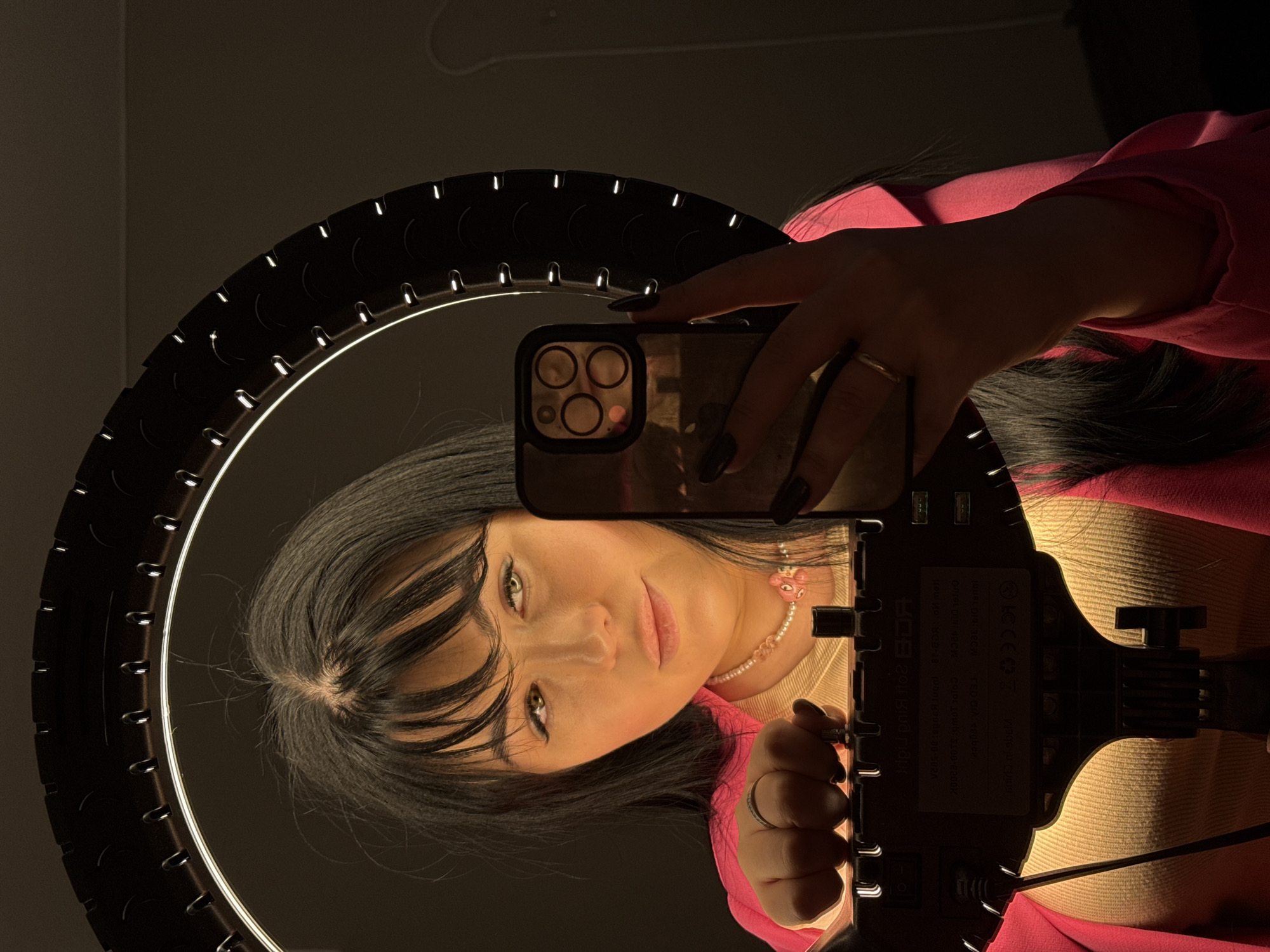 A woman with black hair and bangs is taking a selfie in front of a ring light. She is wearing a pink jacket and a beaded necklace with a pink flower charm. Her phone is in her hand, and the reflection in the phone shows a dark room. The ring light is a black circle with white LEDs around the inner edge. The base of the ring light is visible at the bottom of the frame. The location is not specified in the prompt and is not discernible from the image.
