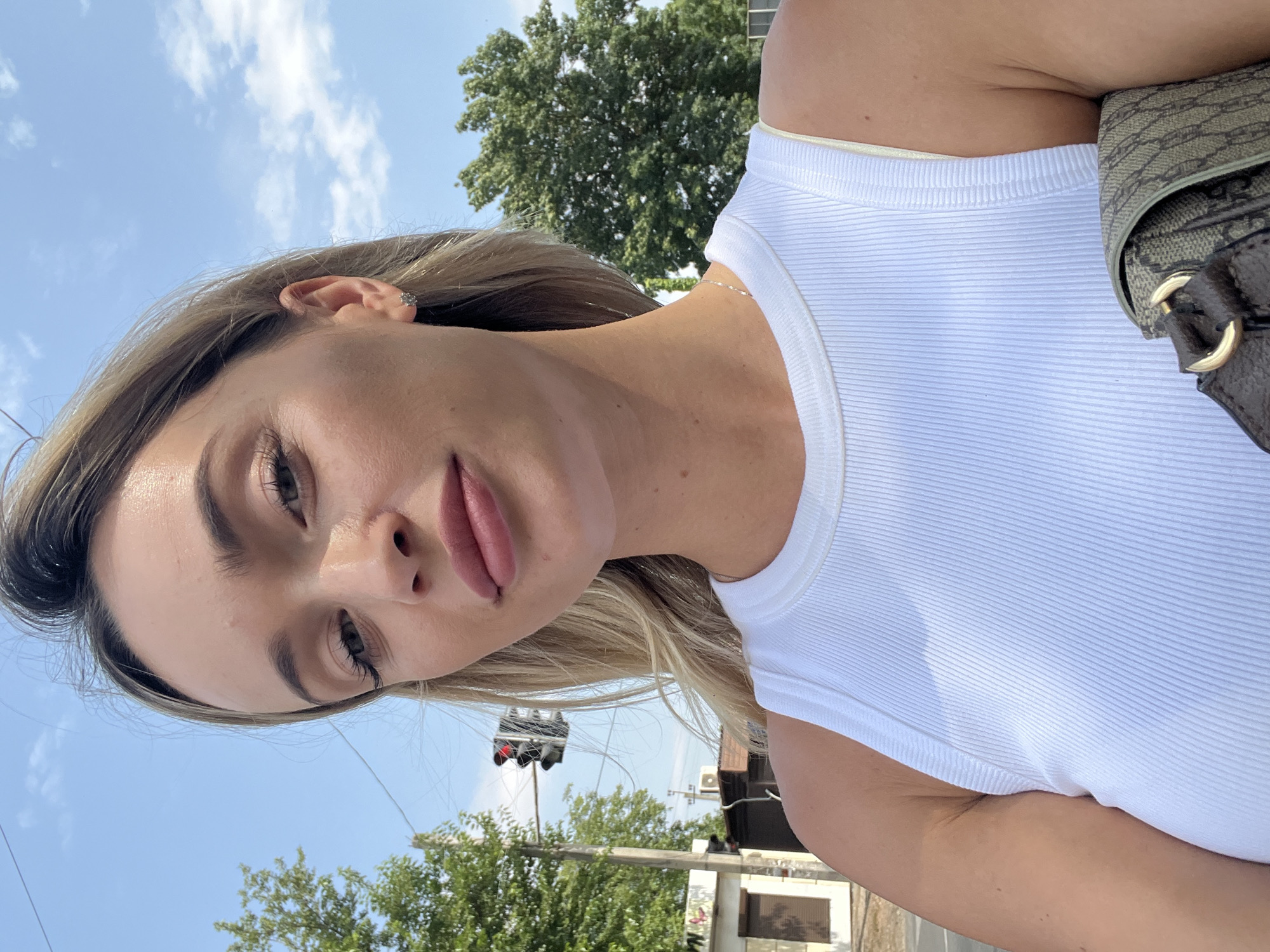 A blonde woman is taking a selfie outdoors under a bright blue sky with scattered clouds. She is wearing a white ribbed tank top and a necklace. A patterned handbag with gold hardware is visible in the lower right corner of the frame. In the background, there is a traffic light with a red light illuminated, trees, and a building. The location is identified as Fontanka, Ukraine.