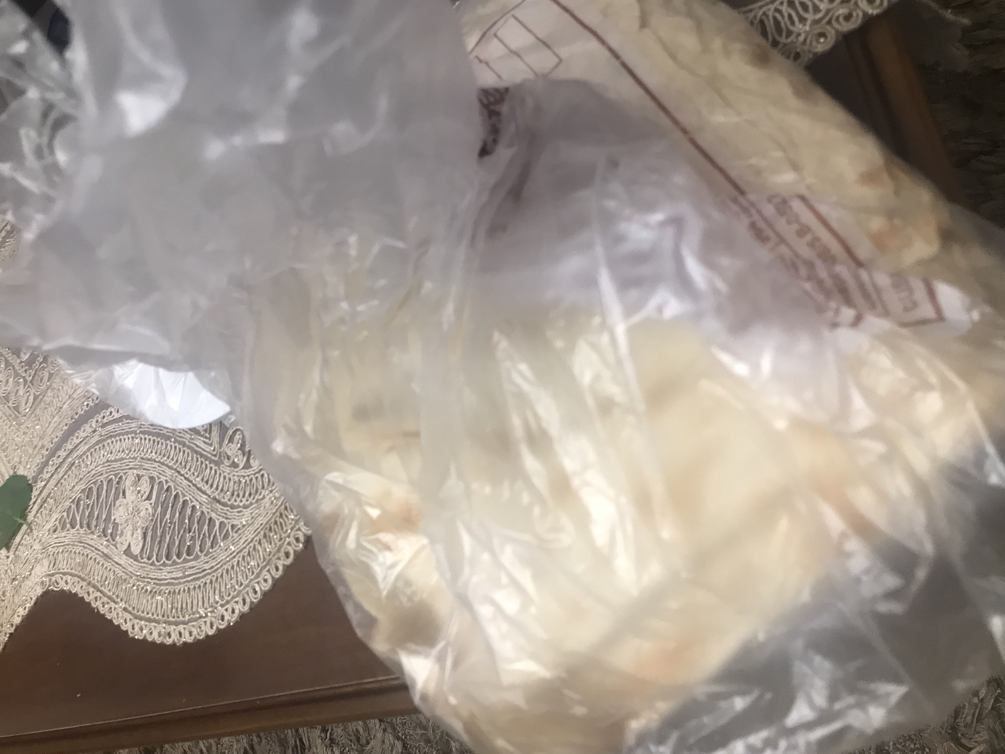 The image shows a clear plastic bag filled with bread or pita-like flatbread. The bag is on a table covered with a beige, lacy tablecloth. Parts of a dark wooden surface are visible beneath the tablecloth. The location is unknown, the image contains no information about Yerevan or Armenia.
