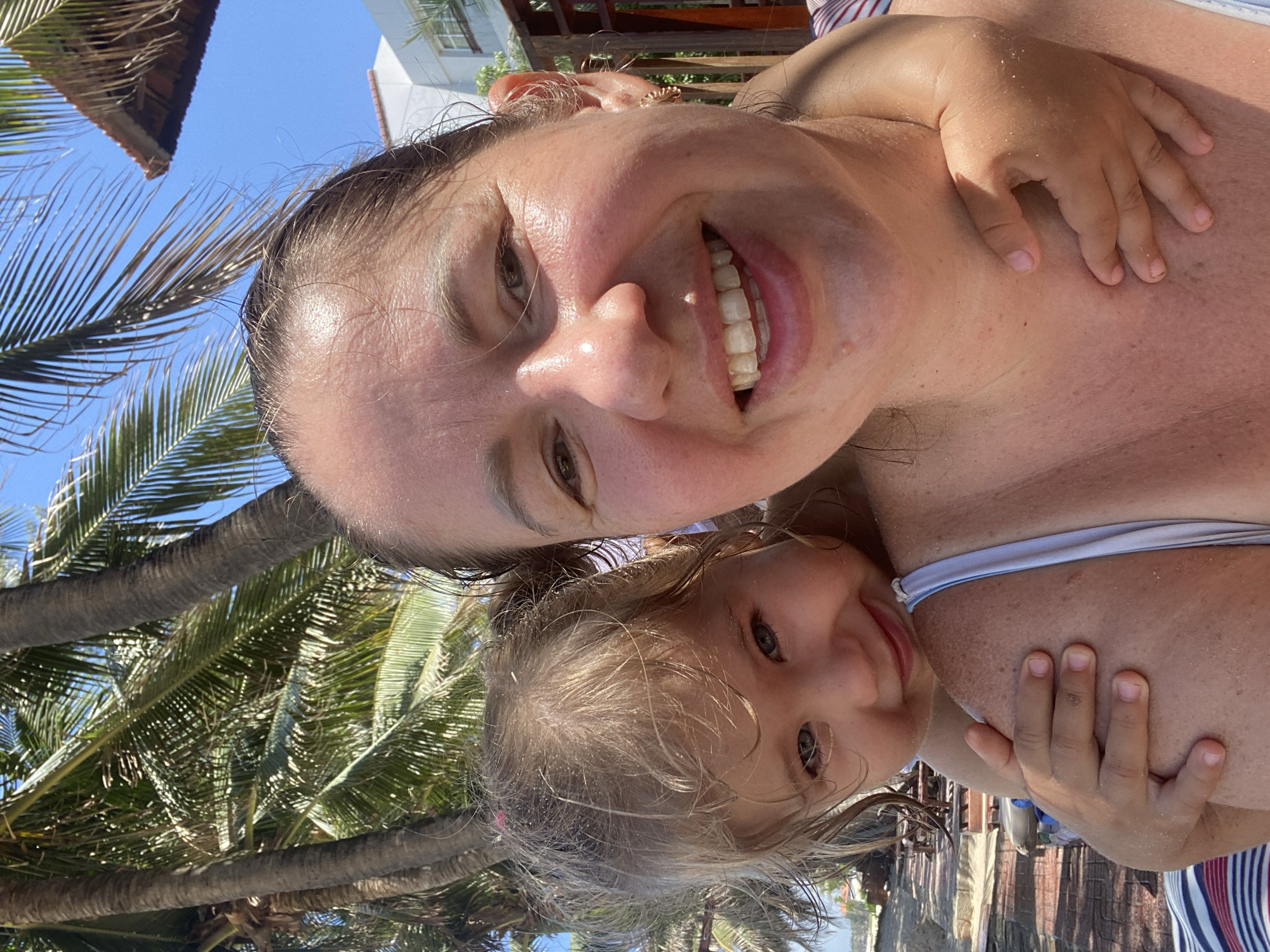 The image is a selfie taken outdoors with a bright blue sky. The subjects are two people, an adult and a young child, possibly mother and daughter, who appear to be on a beach or near palm trees. The adult is in the foreground, smiling widely and showing her teeth. Her head is tilted, and her face is illuminated by sunlight. A child's hand is resting on her chest. The child is also visible in the frame, with blonde hair and a smiling expression, looking towards the camera.
The setting includes lush green palm trees and what appears to be a building with a white facade in the background, suggesting a tropical or beachside resort location. The presence of sand on the skin of the adult and child, along with the palm trees and bright sky, indicates a sunny day. The overall mood conveyed is one of happiness and a relaxed vacation atmosphere. The image seems to capture a moment of shared joy between the two individuals, likely during a holiday. There is no visible text in the image.