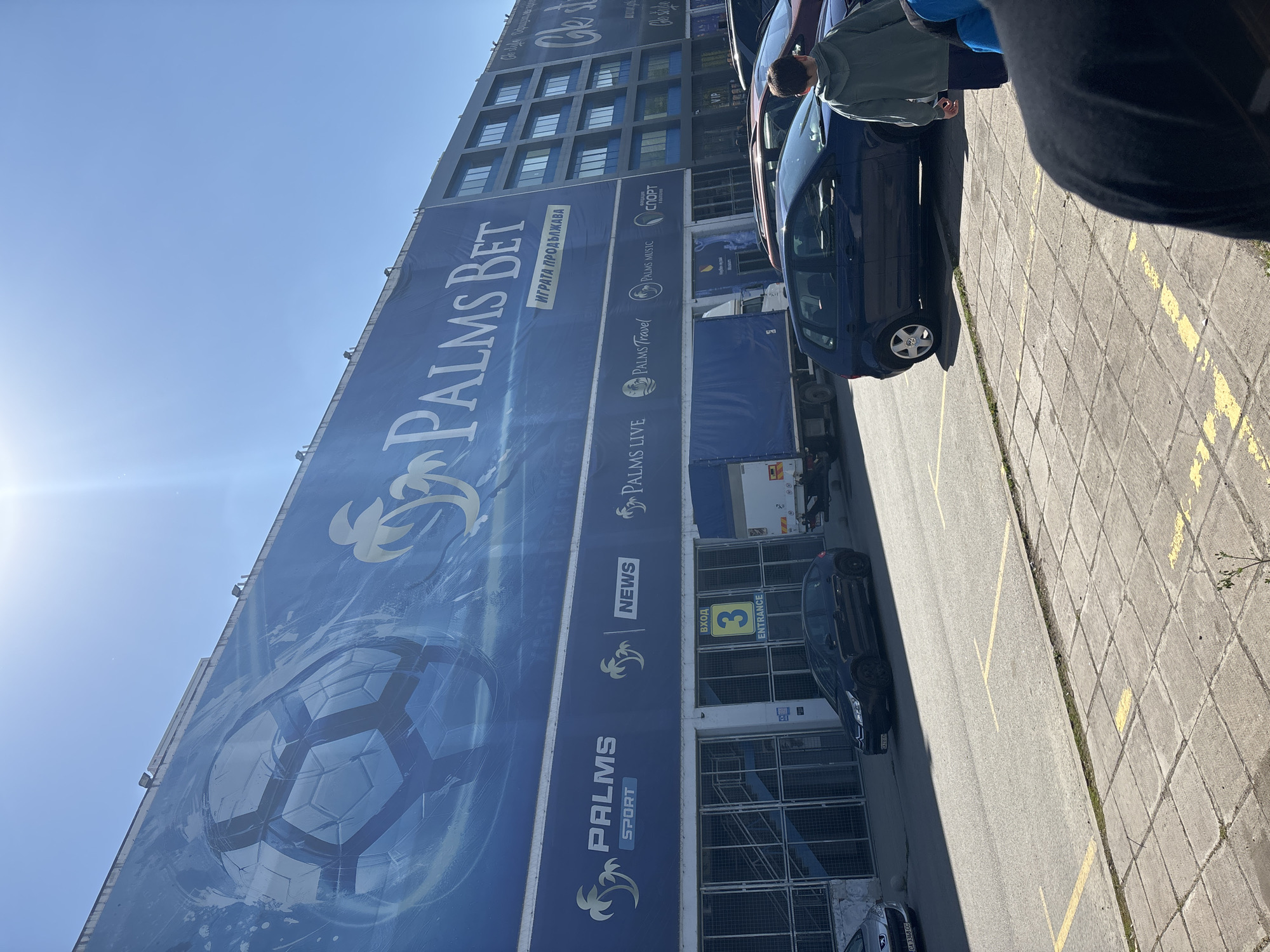 A brightly lit exterior view captures a large commercial building in Sofia, Bulgaria, on a clear, sunny day. The image is oriented vertically, showing a massive blue banner covering most of the building's facade. The sky, a light blue, occupies the upper left portion, with a sun flare visible.
The prominent banner features "PALMS BET" in large, stylized white and gold letters, with the Bulgarian phrase "ИГРАТА ПРОДЪЛЖАВА" (The game continues) directly beneath it. Further down, there's smaller text "ВАРТОТ НОСИ РИСК В" (The value carries risk). Below this, various sub-brands are advertised with their logos: "PALMS LIVE", "PALMS Travel", "PALMS MUSIC", "PALMS SPORT", and "NEWS". A large, abstract graphic resembling a soccer ball or geometric sphere is visible on the lower left of the banner. Near the bottom right, an entrance is marked with a yellow sign reading "ВХОД 3 ENTRANCE".
In the foreground and to the right, a paved parking area with yellow dividing lines is visible. Several vehicles are parked: a dark blue hatchback (likely a Volkswagen, identifiable by its wheel cap) is central, with a person wearing a green jacket and blue pants standing nearby, facing away. A large white truck with a blue tarp-covered trailer is parked closer to the building. Another light-colored car, partially visible, displays a Bulgarian license plate "CA 3368 CC", confirming the Sofia location. Part of a person's leg and shoe is visible in the extreme foreground, partially obscuring the pavement. The scene depicts a typical commercial outdoor area during daytime.