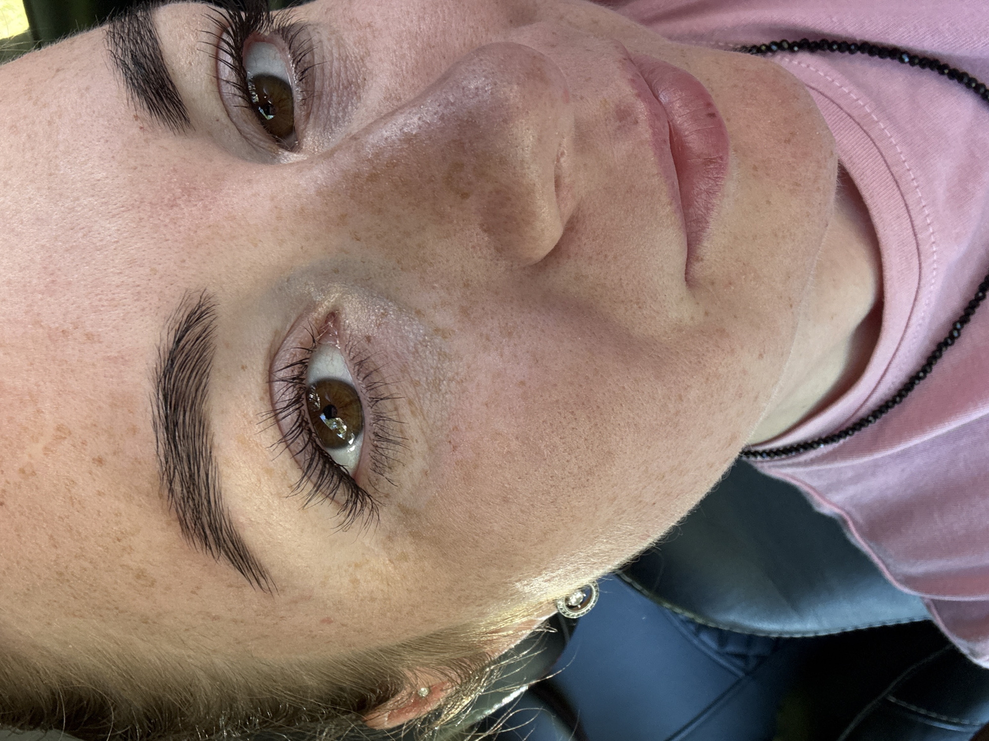 A close-up shot features the face of a fair-skinned woman with dark eyebrows and brown eyes, adorned with long, dark eyelashes. She has numerous freckles across her forehead, cheeks, and nose. The woman is wearing a pink t-shirt and a beaded necklace. A circular earring with a dangling charm is visible on her left earlobe. The background is out of focus, showing a dark-colored surface, likely the interior of a vehicle. The lighting suggests an outdoor setting, possibly during the day.