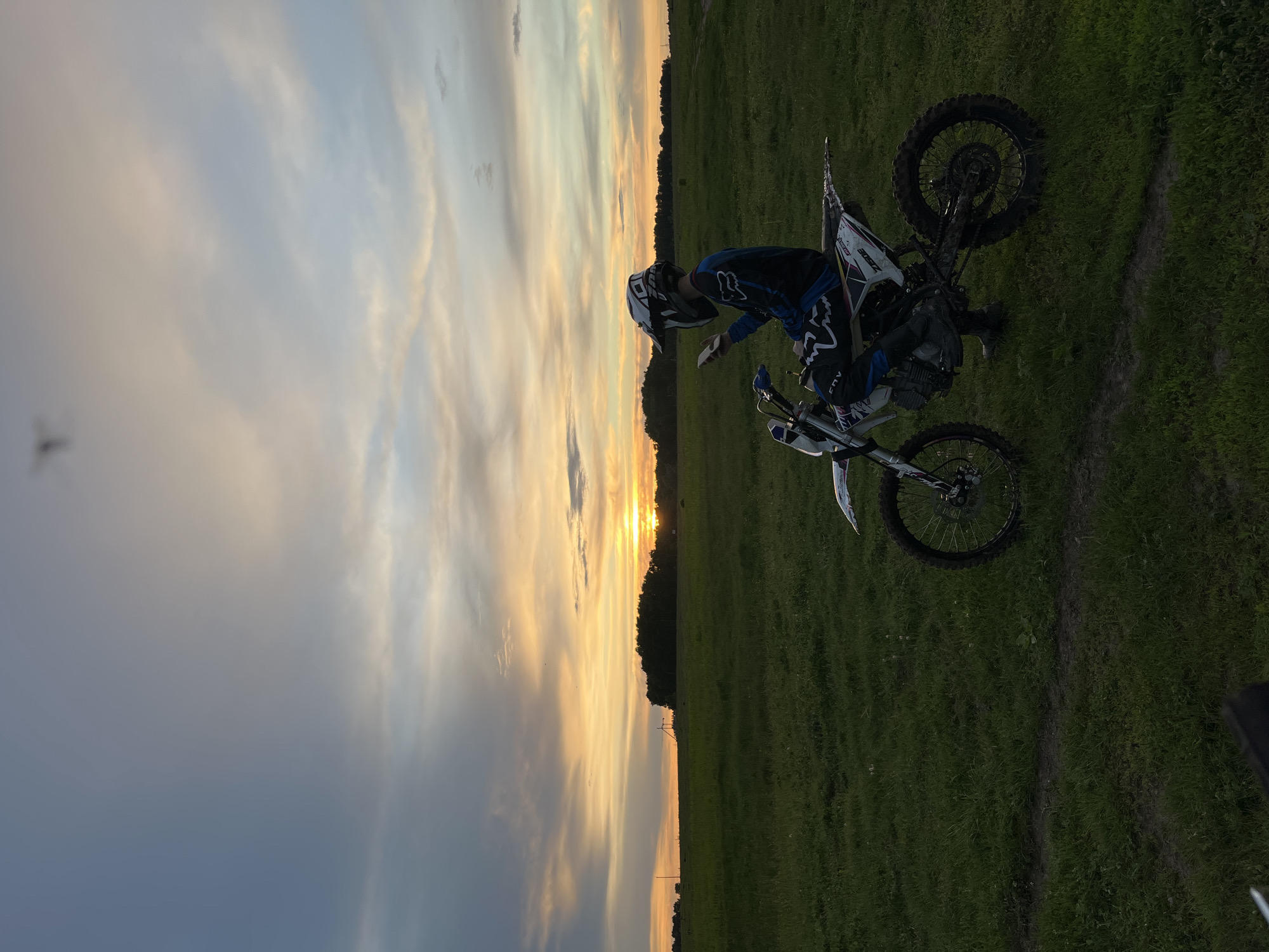 A person in motocross gear, including a helmet and blue and black clothing, is standing next to a dirt bike on a grassy field. The dirt bike is mostly white with black accents. The sky behind them is a gradient of colors, transitioning from a dark blue/grey on the left to a bright orange at the horizon, suggesting it is either sunrise or sunset. A line of trees is visible on the horizon. The location is Новочувашский, Russia.