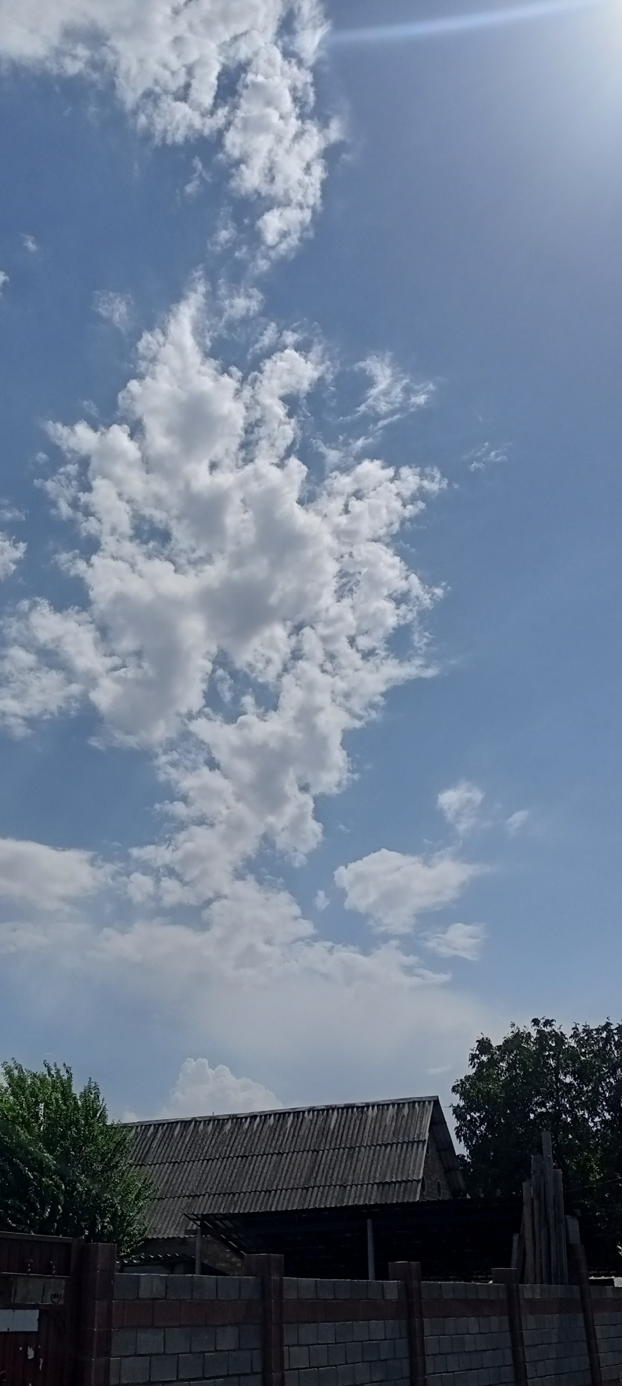 The image depicts a cloudy sky over a rural scene in Sokuluk, Kyrgyzstan. The sky is predominantly blue with scattered white cumulus clouds. A portion of the sun is visible in the upper right corner, creating a slight lens flare. In the foreground, there is a weathered, grey, corrugated roof of a building. To the left of the building, a lush green tree is visible, and to the right, a darker green tree with dense foliage. A fence made of concrete blocks runs across the lower part of the image, with red and grey horizontal bands. Some wooden posts and building materials are stacked behind the fence on the right side of the frame. The overall impression is one of a slightly overcast day in a residential or agricultural area. There are no people or animals visible in the image.