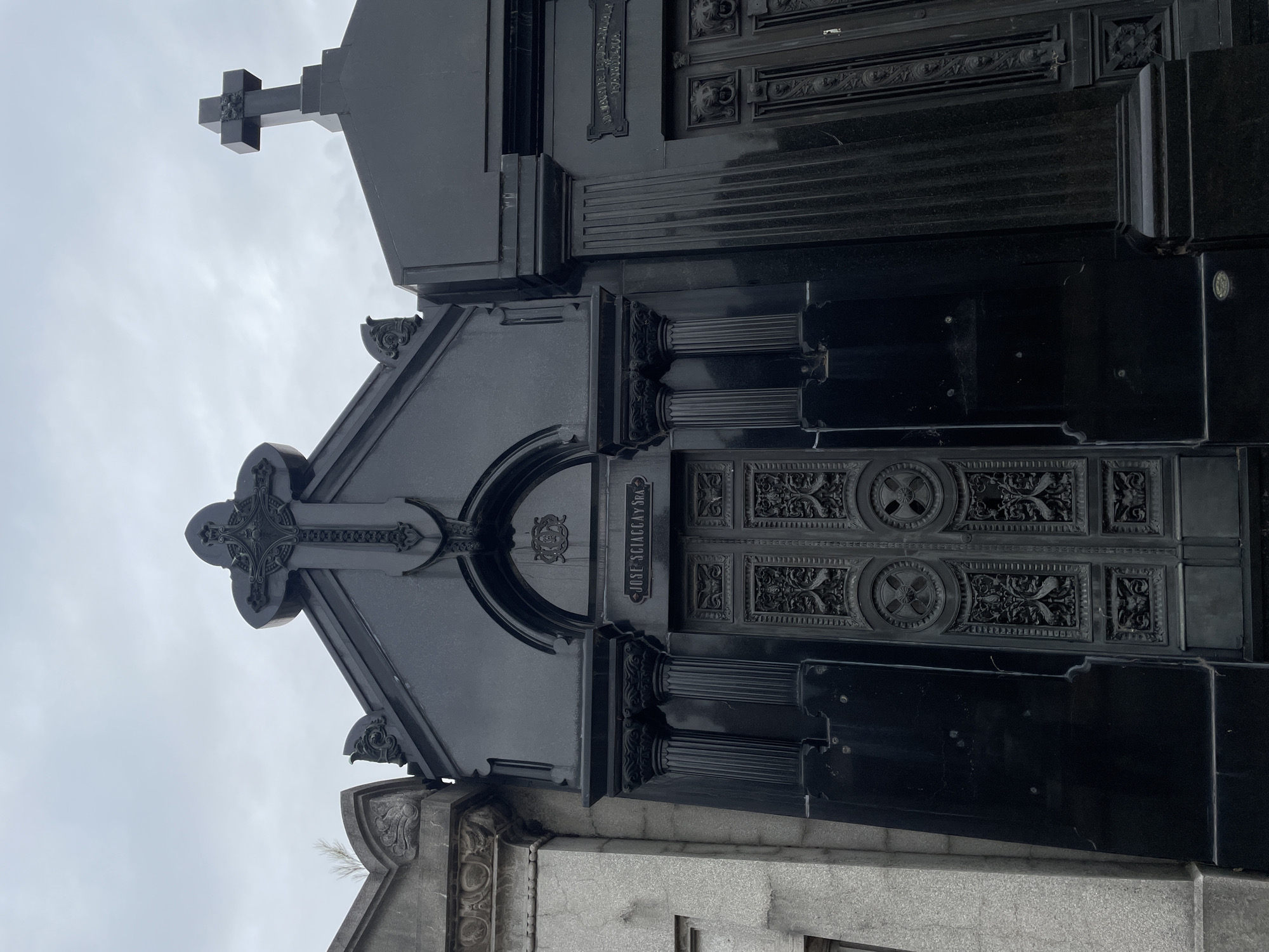 The image displays three ornate mausoleums within Recoleta Cemetery in Buenos Aires, Argentina, under an overcast sky. The central mausoleum is constructed from dark, polished stone or black granite, featuring a prominent gabled roof capped by a large, intricately carved cross adorned with vegetative and geometric patterns. Below the gable, an arched recess houses a pair of heavy, dark metal double doors, which are elaborately decorated with circular medallions containing crosses and intricate scrollwork. Above these doors, an inscription reads "JOSE SCIACCA Y SRA." The entrance is flanked by two fluted columns with Corinthian capitals. To the left, a partial view of an adjacent mausoleum made of lighter grey stone is visible, characterized by a scalloped roofline and carved embellishments. To the right, another dark stone mausoleum is partially shown, topped with a simpler, solid cross and featuring a partially legible plaque reading "SALVADORES DE GUELMAN" with associated dates. No individuals are present in the scene.