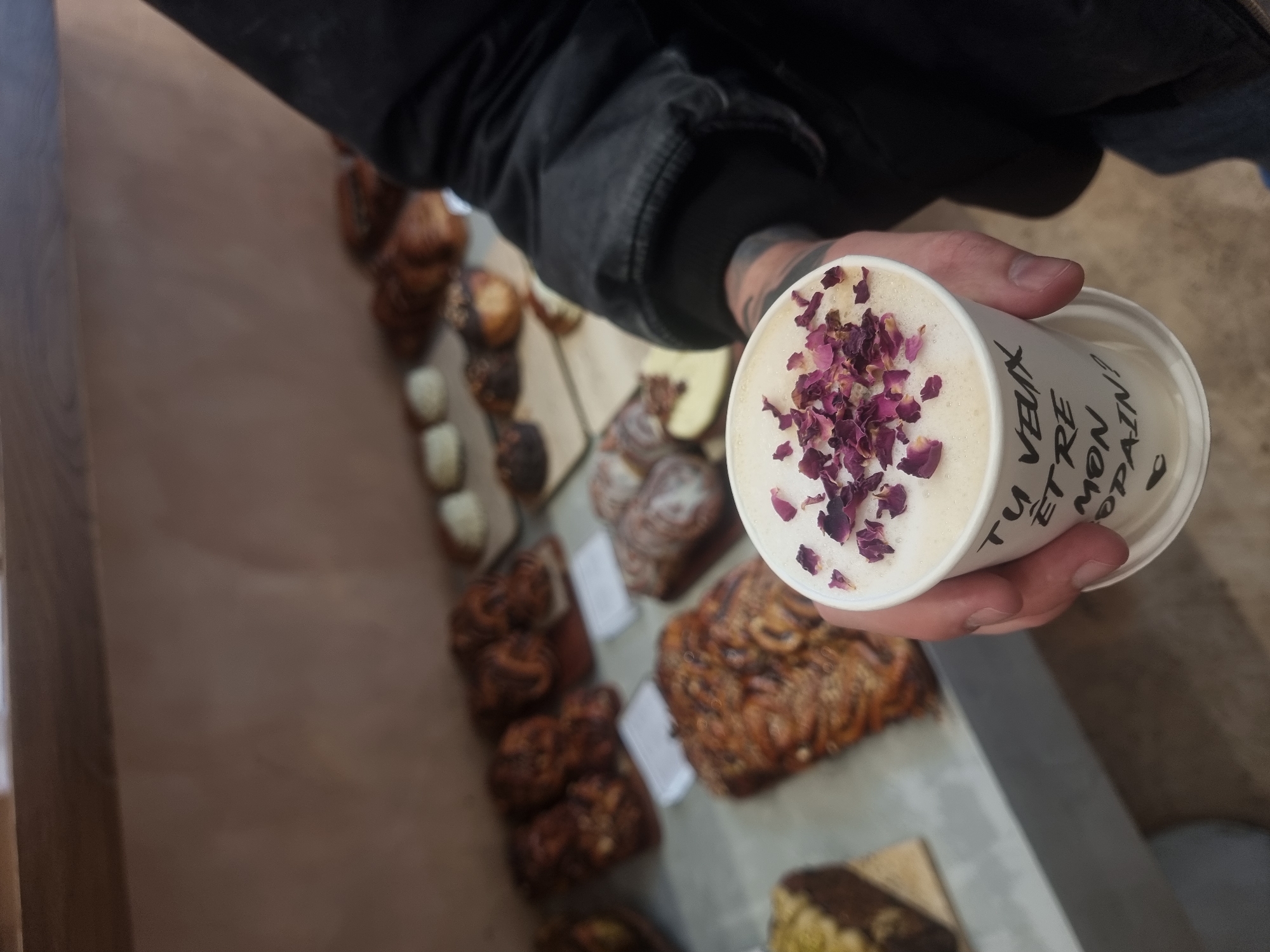 An individual holds a white takeaway cup filled with a beverage topped with pink and purple dried rose petals, likely a rose latte or similar drink. The person's arm is visible, wearing a dark jacket, and a tattoo can be seen on their forearm. On the side of the cup, black handwritten-style text reads "TU VEUX ETRE MON COPAIN?", which translates from French to "Do you want to be my boyfriend?".
The scene is set indoors, likely a bakery or cafe in Paris, France, suggested by the French text and the array of baked goods in the background. Out of focus behind the hand and cup, a display counter showcases various pastries and baked items. These include large, swirled loaves resembling babka, smaller individual buns, some with dark glazes or toppings, and other lighter-colored pastries. The presentation of the food suggests an artisanal establishment. The lighting appears natural and soft, highlighting the beverage and pastries. The image captures a moment of enjoying a unique drink in a charming culinary setting.