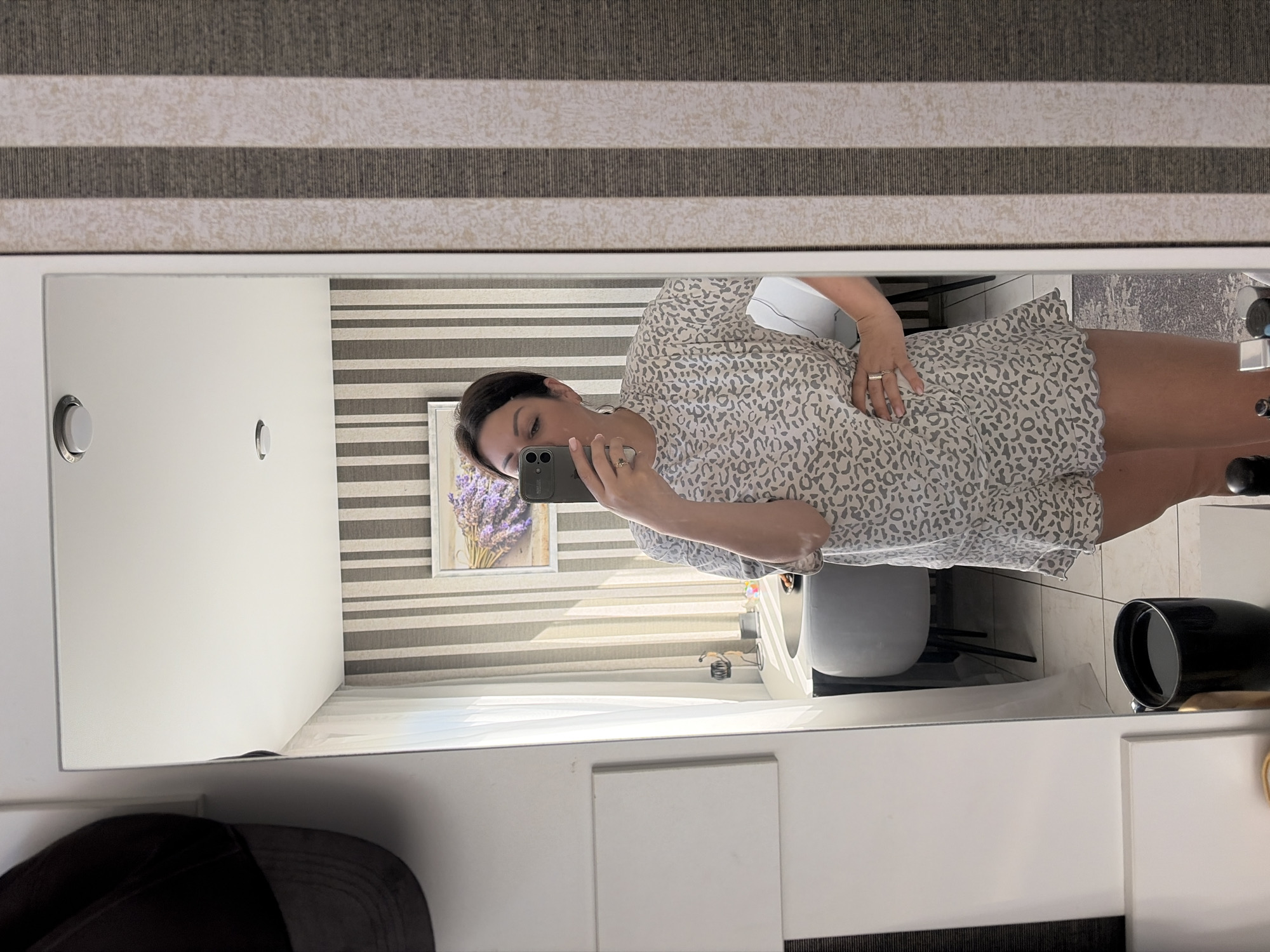 In a well-lit indoor setting, a woman stands in front of a large mirror, taking a selfie. She is wearing a light-colored, short-sleeved pajama set with a subtle leopard-like print. The pajama top appears to be a t-shirt style, while the bottoms are shorts with a scalloped hem. Her hair is pulled back, and she is wearing jewelry, including rings on her fingers.
The mirror reflects the room behind her, which features a striped wallpaper in shades of beige and dark gray. A framed picture of lavender flowers hangs on the wall. To the left of the mirror, a black baseball cap is visible hanging on a hook. In the reflection, a modern-looking desk with a gray chair is seen, along with some items on the desk. On a surface in the foreground, a black mug filled with a dark liquid is present. The floor appears to be tiled.
The scene is not specifically located in Sochi, Russia. The image depicts a residential or hotel room interior.