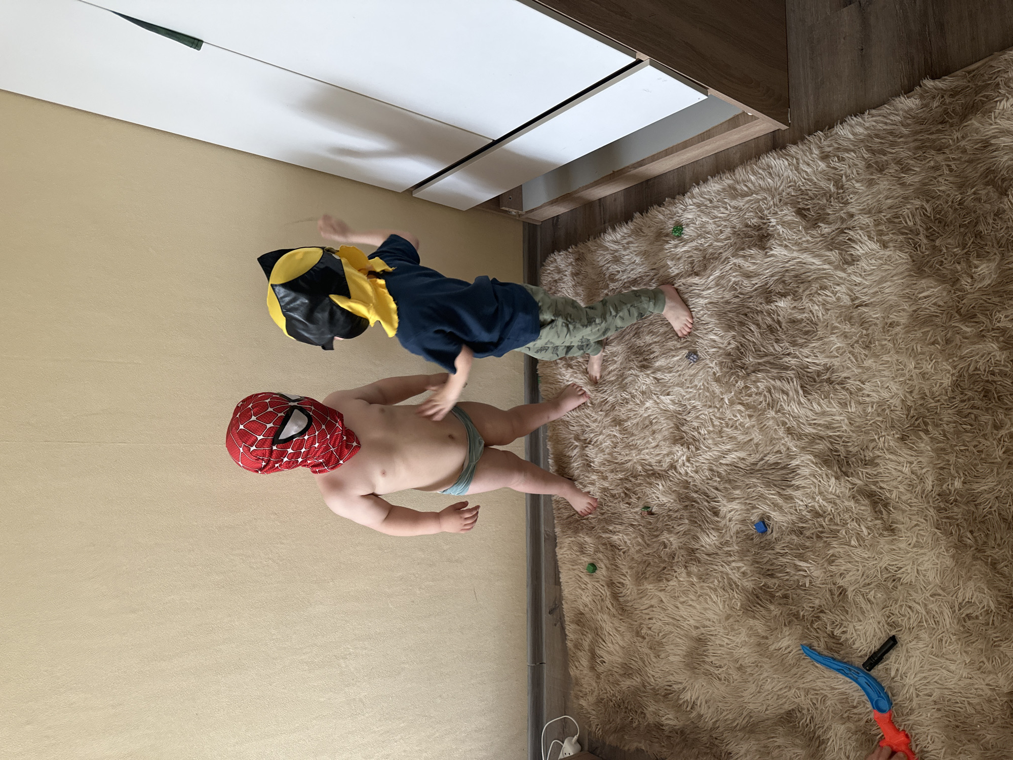 The image, taken in the city of Chaikovsky, Russia, depicts two young children playing indoors. On the left, a child wearing a red Spiderman mask that covers the entire head, and light blue briefs, stands barefoot. To the right, another child, wearing a yellow and black mask, a dark blue short-sleeved t-shirt, and camouflage trousers, stands barefoot, extending their right hand towards the first child's torso. Both children are positioned on a shaggy, light brown area rug that covers most of the floor. A dark wood-patterned laminate floor is visible around the rug's edges, particularly on the left and right sides. The background features a plain, textured light beige wall. On the far right, a tall, white wardrobe with a light wood frame is visible. Several small plastic toys, including a blue and orange toy sword, a black cylindrical object, and green and blue blocks, are scattered on the rug. A white electrical outlet and cord are visible on the floor against the wall in the lower-left corner.
