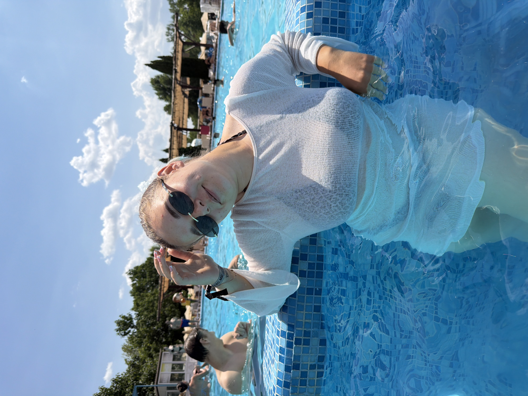 A young woman, wearing sunglasses and a sheer white cover-up over a bikini, leans against the tiled edge of a swimming pool. She is partially submerged in the bright blue water, with her right arm resting on the pool edge. Her left hand is touching her hair. The background shows other people swimming in the pool and lounging in the distance, with trees and a covered seating area visible under a clear blue sky with scattered clouds. The image appears to be taken outdoors at a public swimming pool, possibly in Sociteni, Moldova, given the context.