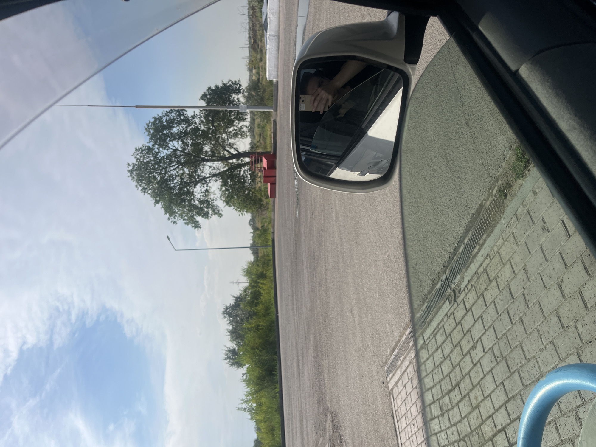 The image captures an outdoor scene, likely a paved area with some greenery and structures in the background, seen from the perspective of being inside a vehicle. A side mirror of the vehicle reflects a person holding a phone, seemingly taking a selfie. The environment includes a large tree, a street lamp, and what appears to be a low wall or structure in the middle ground. The ground is paved with interlocking stones in the foreground and asphalt further back. The sky is partly cloudy with blue patches. The location is identified as Aksu, Kazakhstan.