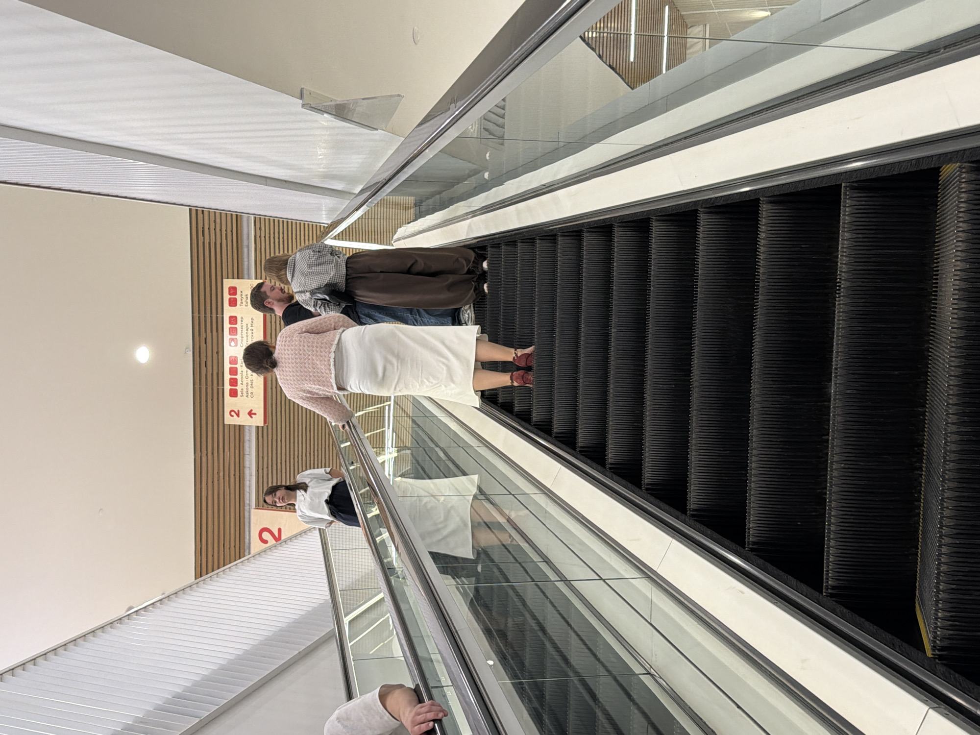 Four individuals are ascending an escalator in a modern indoor environment, likely a shopping mall or public building. The escalator is made of black metal with transparent glass railings on either side. In the foreground, the escalator steps are clearly visible.
The individuals, from left to right, are:
1. A person whose left arm and shoulder are visible, resting on the escalator handrail. They are wearing a light-colored shirt.
2. A young woman with dark hair, wearing a white t-shirt and dark pants. She is looking forward.
3. A woman with her back to the camera, wearing a light pink cardigan over a white skirt and red shoes. She is also looking forward.
4. A man wearing a dark shirt and blue jeans, speaking with a woman to his right.
5. A woman wearing a checkered shirt and brown wide-leg pants, standing next to the man.
Above and behind the individuals, there is a sign with the number "2" and directional arrows, indicating a floor or section. The background features wooden paneling and white architectural elements, suggesting a contemporary design. The lighting appears to be artificial, consistent with an indoor setting. The environment is clean and well-maintained.
The image is captured from a slightly low angle, looking up the escalator. The overall scene depicts everyday movement within a public space. The presence of the "2" sign and the specific signage in the background, including Cyrillic text ("Торговый центр", "Спортмастер"), strongly suggest the location is a shopping center in Kazan, Russia.