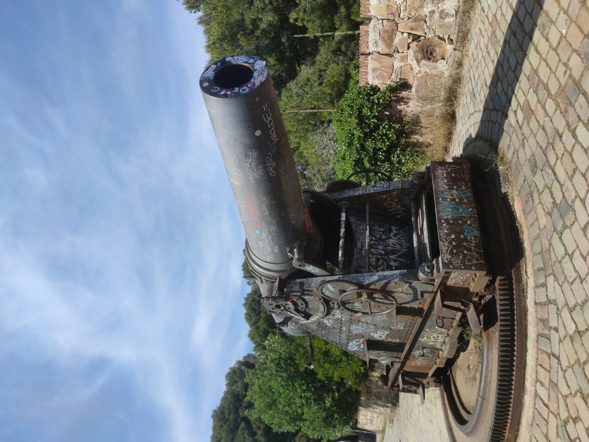 The image shows a large, antique cannon covered in graffiti and stickers. It is situated outdoors on a brick-paved surface, next to a stone wall with vegetation growing around it. Trees are visible in the background, and the sky is partly cloudy. The cannon appears to be a historical artifact, possibly on display in a park or historical site. Given the context of Barcelona, Spain, it is likely that the cannon is located in one of the city's historical sites, potentially Montjuïc Castle or a similar location.