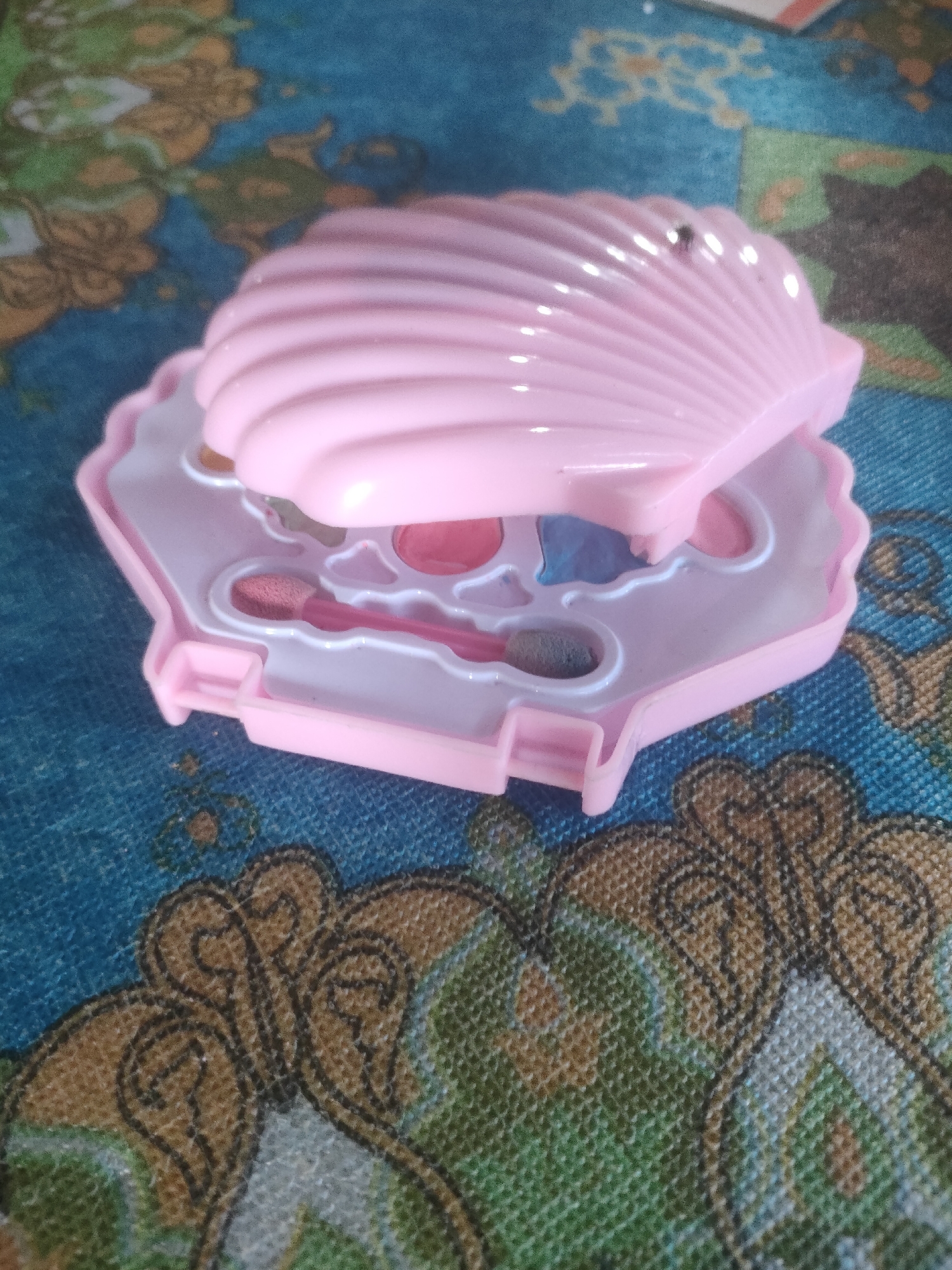 A close-up shot features a pink, shell-shaped children's makeup compact. The compact is open, revealing compartments for eyeshadows and brushes. The exterior of the compact has a ribbed, seashell design. It rests on a patterned blue and green surface, possibly a rug or tablecloth, with intricate brown and black swirling designs. The lighting suggests an indoor environment. No specific location within the city of Padang Dalom, Indonesia is identifiable from the image.