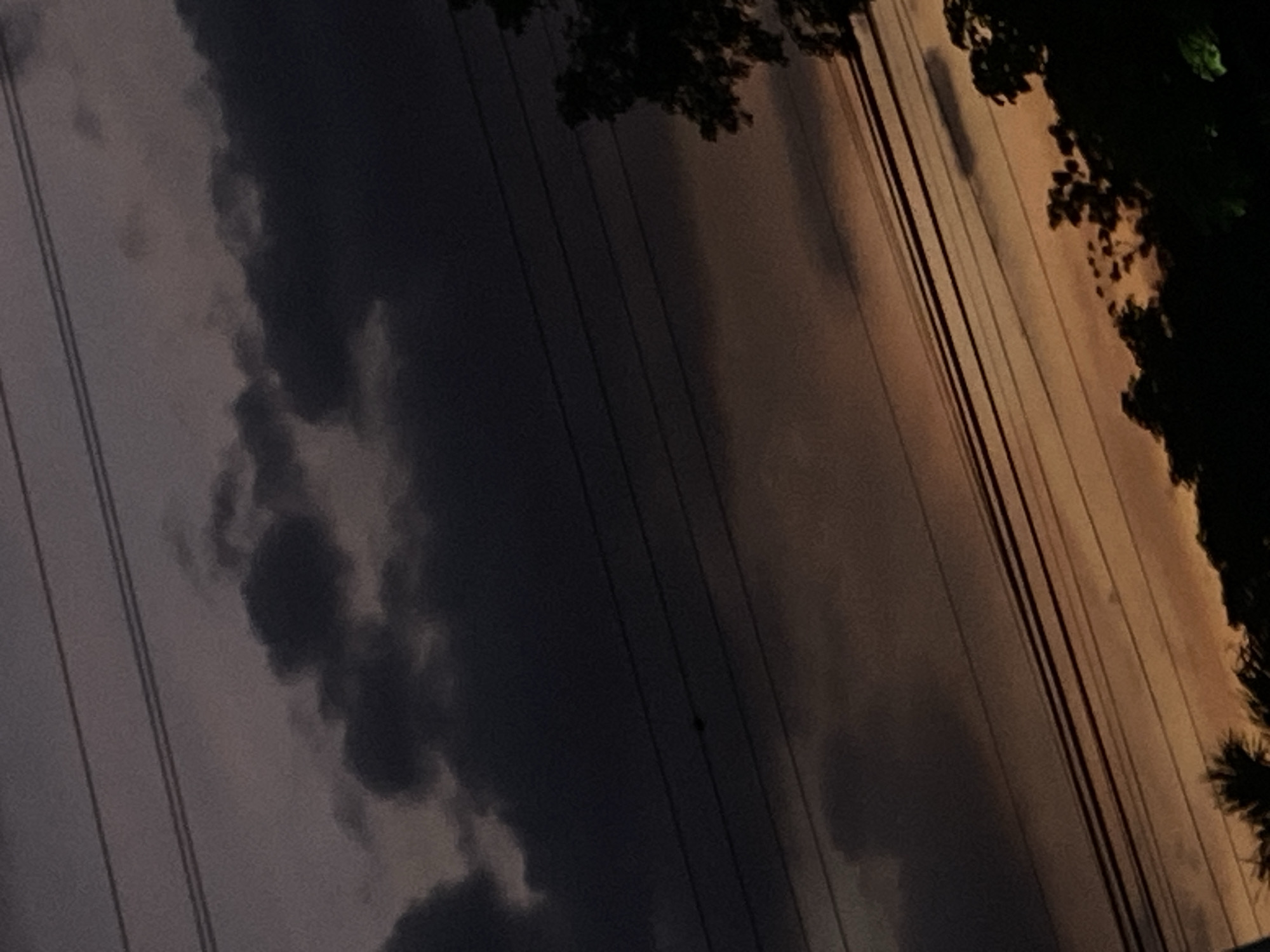 A low-angle, medium shot captures a dramatic dusky sky with an array of dark, heavy clouds dominating the upper portion of the frame. Thin, parallel power lines crisscross the scene horizontally. In the lower half of the image, the sky transitions to a warmer, muted orange hue, suggesting the last vestiges of daylight. Silhouetted against the fading light are the dark shapes of trees and foliage along the bottom edge of the frame, hinting at a natural or semi-rural setting within Tinaquillo, Venezuela. The overall mood is somber and atmospheric, characteristic of an approaching evening or perhaps a storm.