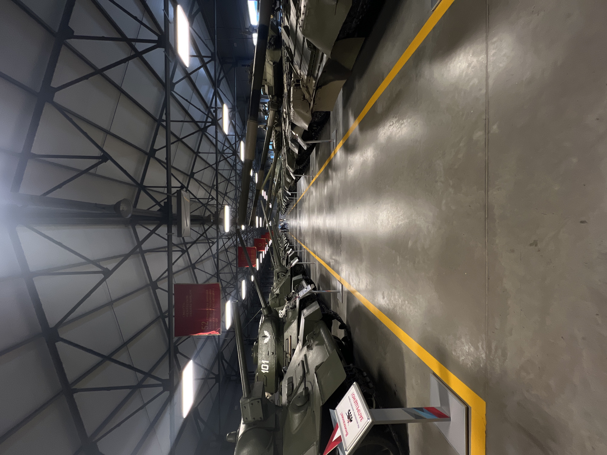 This media file captures an indoor exhibition of military vehicles, likely tanks, within a large, industrial-style building. The scene is dominated by the vehicles, which are parked in a long, orderly line. A bright yellow line runs along the floor, demarcating a walkway or safety zone. The ceiling features a complex metal support structure and is illuminated by fluorescent lighting. Red banners with Cyrillic text are visible hanging from the ceiling.
The visible text includes "Внимание!" (Attention!), "ЗАПРЕЩЕНО!" (Forbidden!), and phrases related to historical events or military units, such as "СМЕРТЬ НЕМЕЦКИМ ЗАХВАТЧИКАМ" (Death to the German invaders), and "ГВАРДИ СКАН ТАНКОВАЯ ФАСТОВСКАЯ АСНОЗНАМЕШАЯ БРИГАДА" (Guards Tank Fastov Red Banner Bogdan Khmelnitsky Brigade). Numbered markings like "52" and "101" are present on the vehicles. One of the tanks has a white marking with a leaf emblem and "101". Another vehicle has a white sign with Cyrillic text and a red and blue striped emblem.
The overall atmosphere suggests a museum or a military exhibition dedicated to historical armored vehicles. The lighting and reflections on the polished floor indicate an indoor, well-lit environment, likely during the daytime. No people are prominently visible in the shot, focusing the attention solely on the preserved military hardware. The image is oriented vertically.