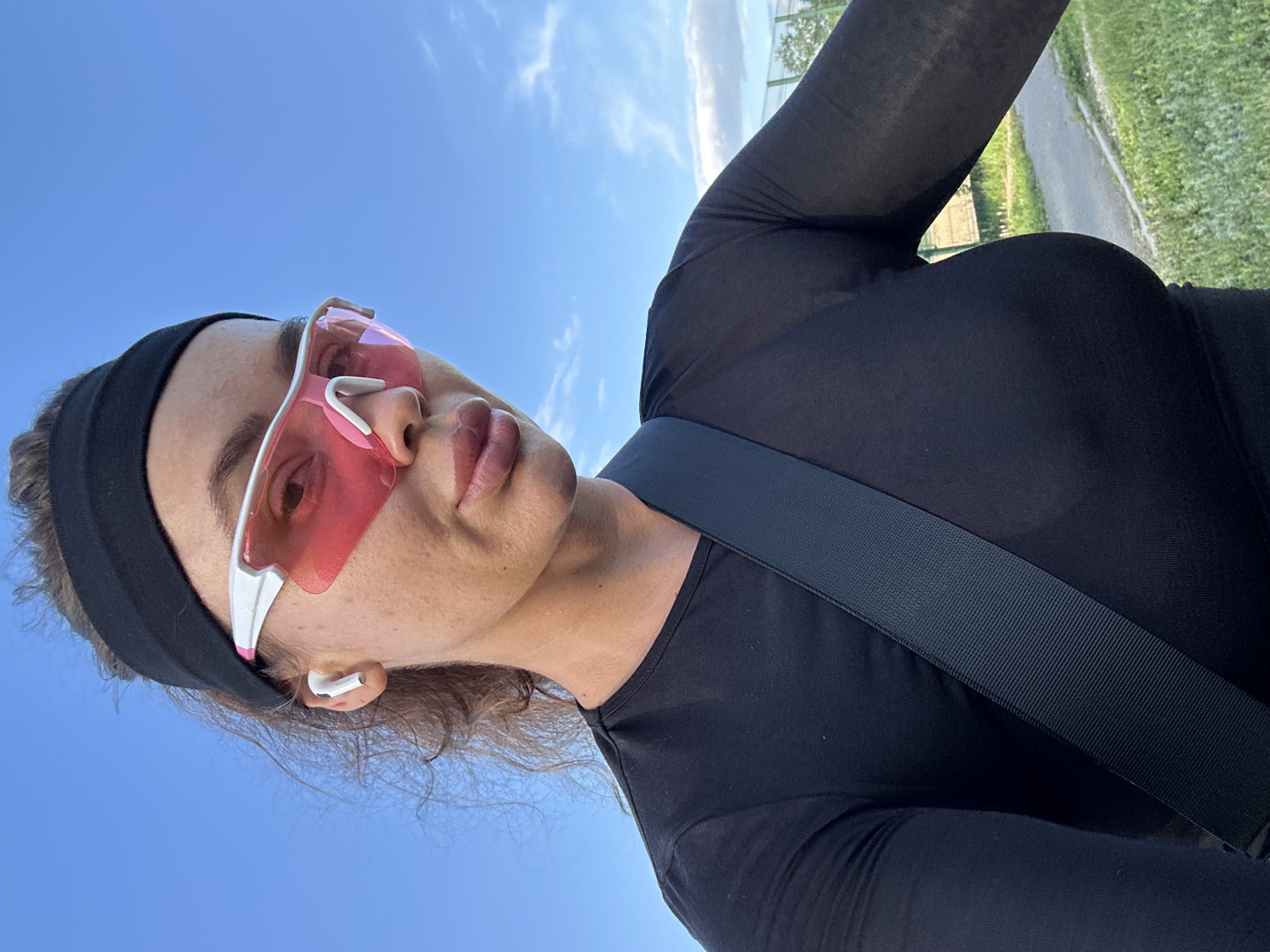 An outdoor, selfie-style, medium shot shows a woman with tanned skin wearing a black athletic top, a black headband, and pink-tinted athletic glasses. She has a dark strap crossing her chest over her top. She is wearing white earbuds. The background is a clear blue sky with some clouds. The location is not specified in the image or context.