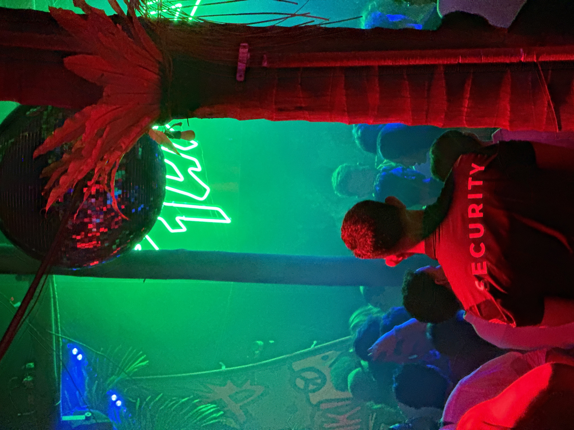 A dimly lit club in North Kuta, Indonesia, is filled with partygoers. The lighting is predominantly green and red. A disco ball adorned with decorative red feathers hangs from a wooden structure, illuminated by the colorful lights. A neon sign is visible in the background. A person wearing a shirt labeled "SECURITY" is seen amid the crowd. The environment is filled with people; several faces are visible in the crowd. A banner bearing graffiti-style art is partially visible in the background.