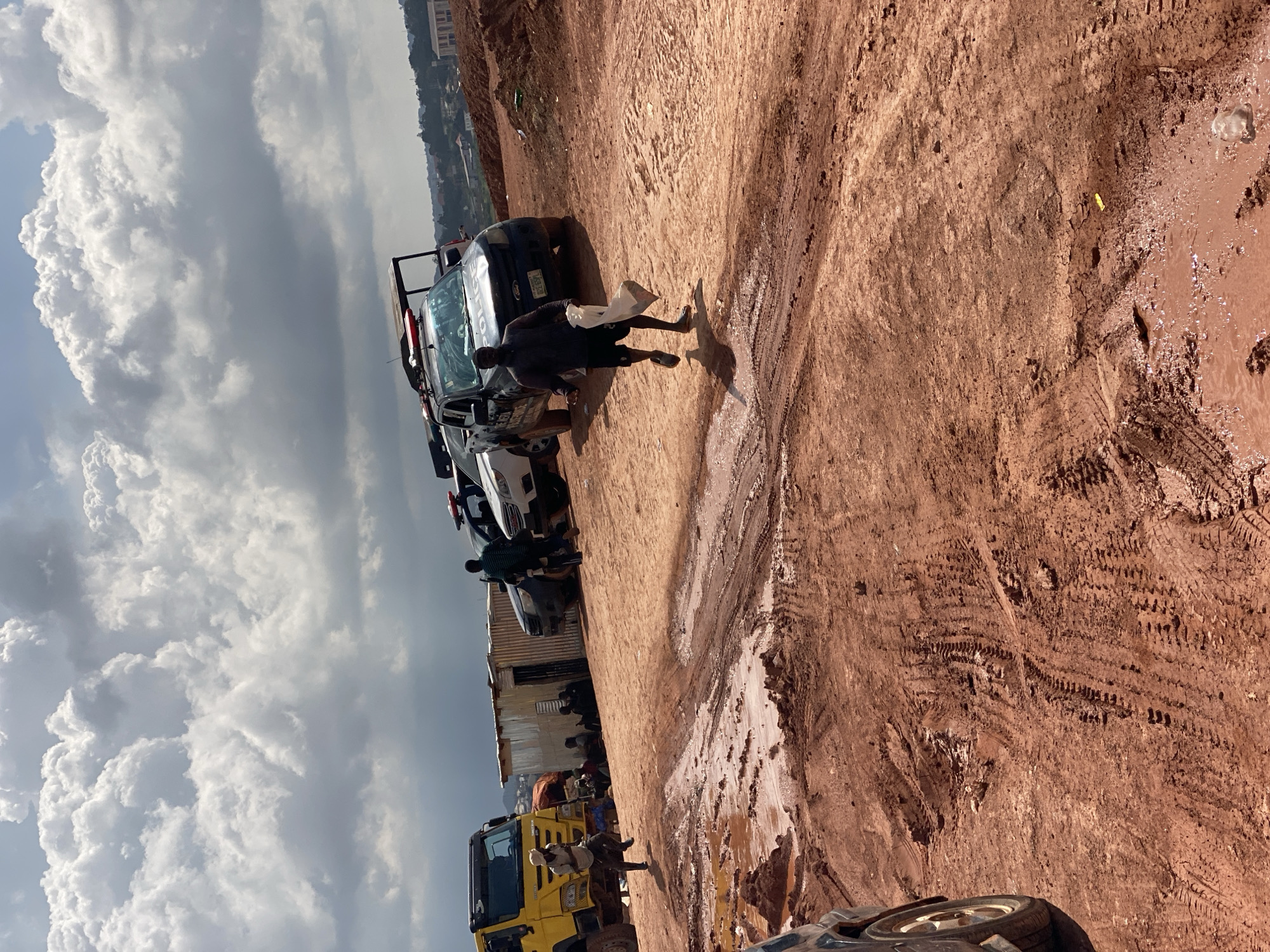 In Reifild-Ist, Nigeria, a scene unfolds under a dramatic sky filled with large, puffy white clouds against a hazy blue backdrop. The ground is a mix of red, muddy earth, marked with tire tracks, and patches of standing water, indicating recent rainfall.
Several vehicles are present, including a police car with visible markings and red and blue lights, suggesting official presence. A yellow truck is also visible, as is another car further down the line.
People are scattered throughout the scene. One man, wearing dark shorts and a dark shirt, walks carrying a white plastic bag. Another man, in a patterned shirt, stands near the police car. Several other individuals are seen near a corrugated metal structure, possibly a makeshift building or shelter. A person in a light-colored shirt appears to be standing on the yellow truck.
The activity appears to be a gathering or a situation involving the police. The presence of multiple vehicles and people on what seems to be an unpaved road in a somewhat undeveloped area suggests a potential incident, a roadside stop, or a community event. The lighting suggests it is daytime, likely mid-morning or afternoon, given the bright sky and shadows. The muddy terrain hints at a tropical climate with a wet season.