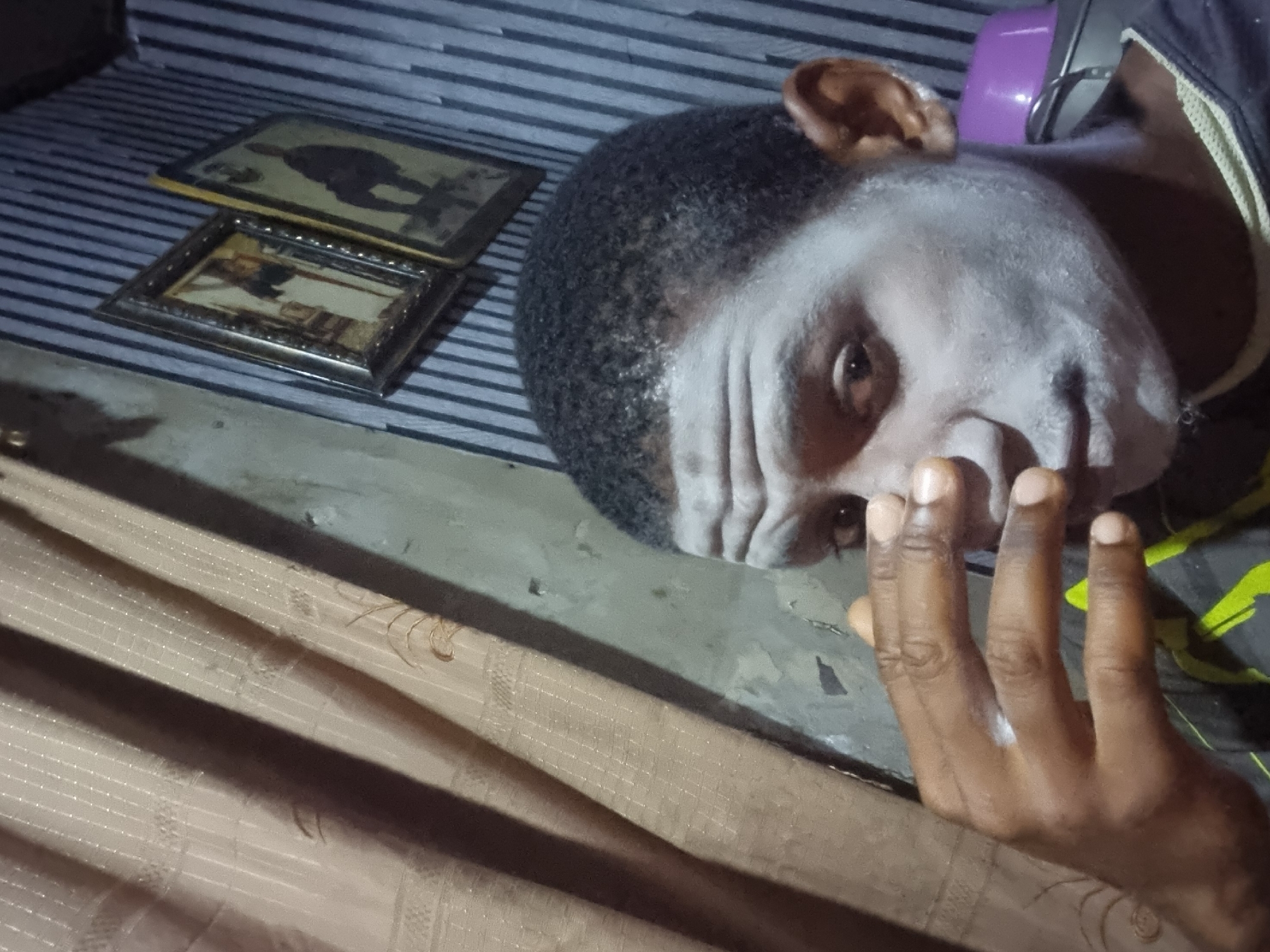 An indoor, dim-lit image features a young adult male, likely in Onitsha, Nigeria, lying on a floor or low surface. His face is notably adorned with white paint or powder across his forehead, cheeks, and nose. He has short, dark, curly hair and looks directly towards the viewer with a neutral, contemplative expression. His right hand is raised, partially obscuring his lower face.
Behind his head, a purple object, possibly headphones, is visible. To the left, two framed pictures are stacked on a striped surface, which could be a bed or mat. The top frame depicts a dark silhouette, while the smaller, lower frame showcases what appears to be a traditional or religious painting with an ornate border. The foreground is occupied by a light brown, subtly textured fabric, possibly a curtain or blanket. The overall subdued lighting suggests the image was taken in the evening or in a poorly illuminated room. No text is visible in the image. The white facial application hints at a potential cultural performance, ritual, or artistic expression common in the region.