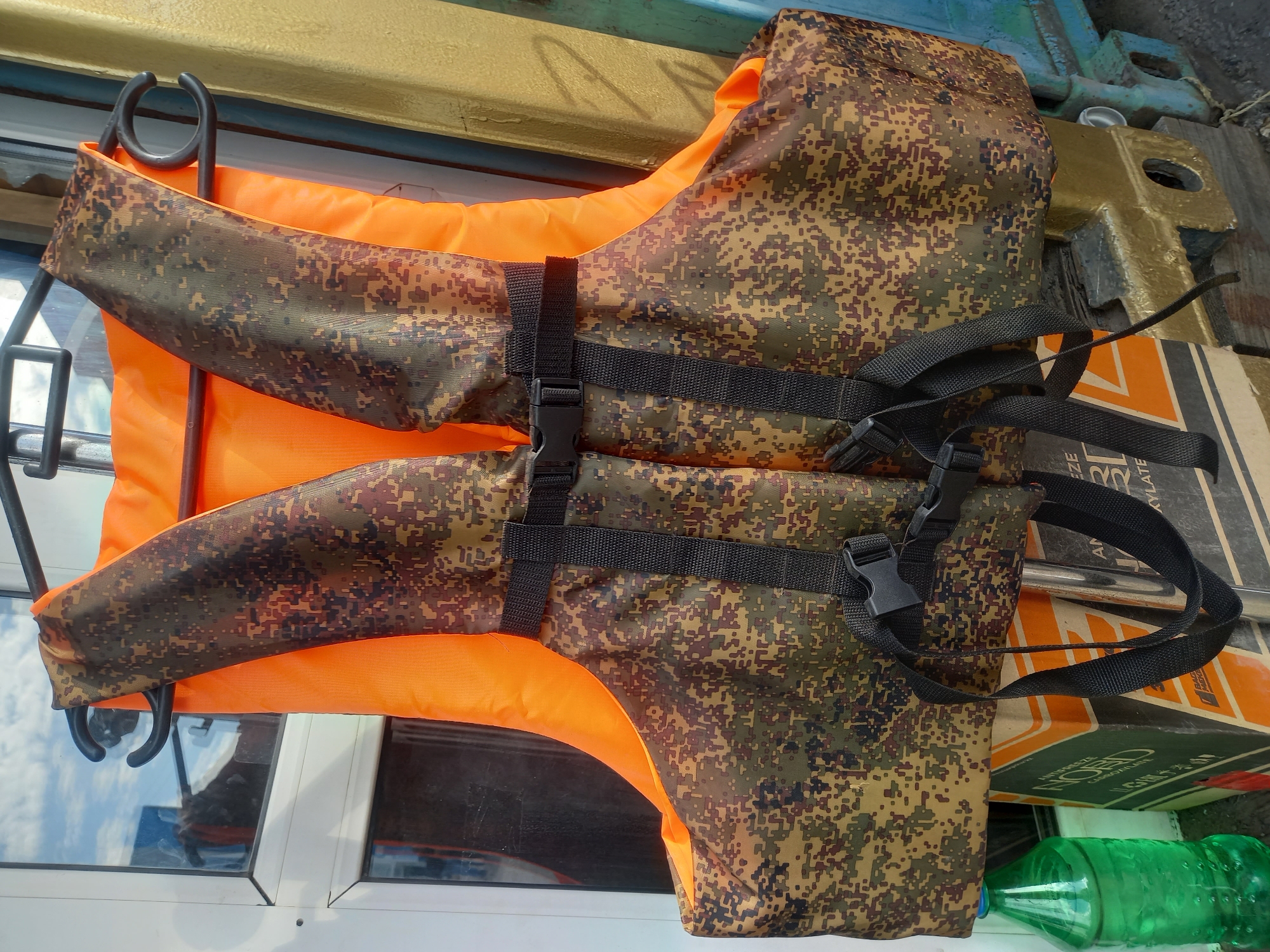 A life jacket with a camouflage pattern and orange interior rests on a surface next to boxes and a green plastic bottle. The life jacket has black straps with buckles. The scene is likely indoors or on a boat, suggested by the surrounding objects and the reflective surface nearby.