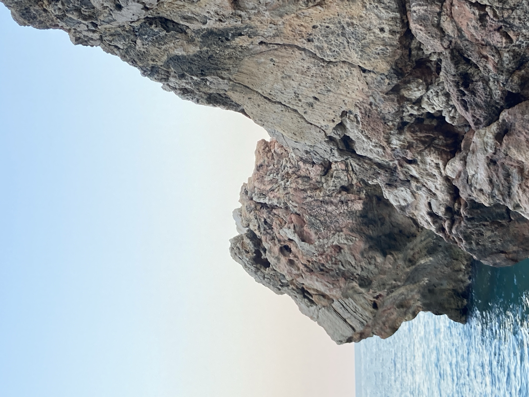 This image captures a close-up view of weathered rock formations jutting out into the sea. The rocks display a rough, textured surface with variations in color, ranging from light tan to reddish-brown and darker gray, indicating mineral deposits and erosion. Some areas of the rock show distinct layers and fissures, suggesting geological age. The sea, a deep blue-green, laps at the base of the rocks, with small waves creating a gentle ripple effect. The sky above is a pale, clear blue, fading to a lighter hue near the horizon, suggesting either early morning or late afternoon. The scene is natural and serene, with no human presence or artificial elements visible. This type of coastal geology is characteristic of the coastline around Kemer, Turkey, a region known for its dramatic cliffs and Mediterranean waters.