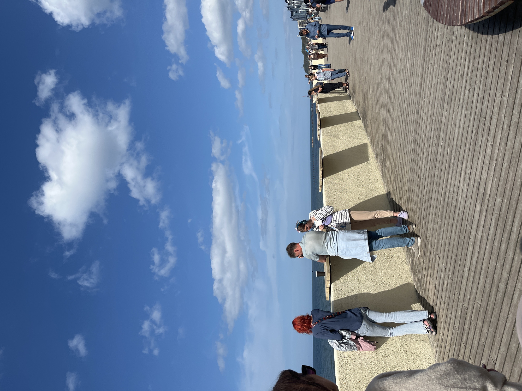 The image depicts a boardwalk along the Baltic Sea in Svetlogorsk, Russia, on a clear, sunny day. Several people are visible on the boardwalk, some walking, others standing and conversing near a low wall overlooking the water. The boardwalk is constructed of wooden planks, and a concrete barrier with a rough, textured surface runs along the edge, separating the walkway from the sea. The sea itself is a deep blue, meeting a bright blue sky dotted with white, fluffy clouds. In the background, buildings can be seen along the coastline.
Specifically, a woman with bright red hair is standing on the left, facing the sea, leaning against the barrier. Next to her, a couple is engaged in conversation at a small wooden table. The man is wearing a light blue shirt and a light blue apron or skirt, and the woman is in a striped top and beige pants. Further down the boardwalk, more individuals are strolling, including a man in a blue shirt and jeans, and a group of people standing together. The overall scene conveys a relaxed atmosphere of people enjoying the seaside promenade.