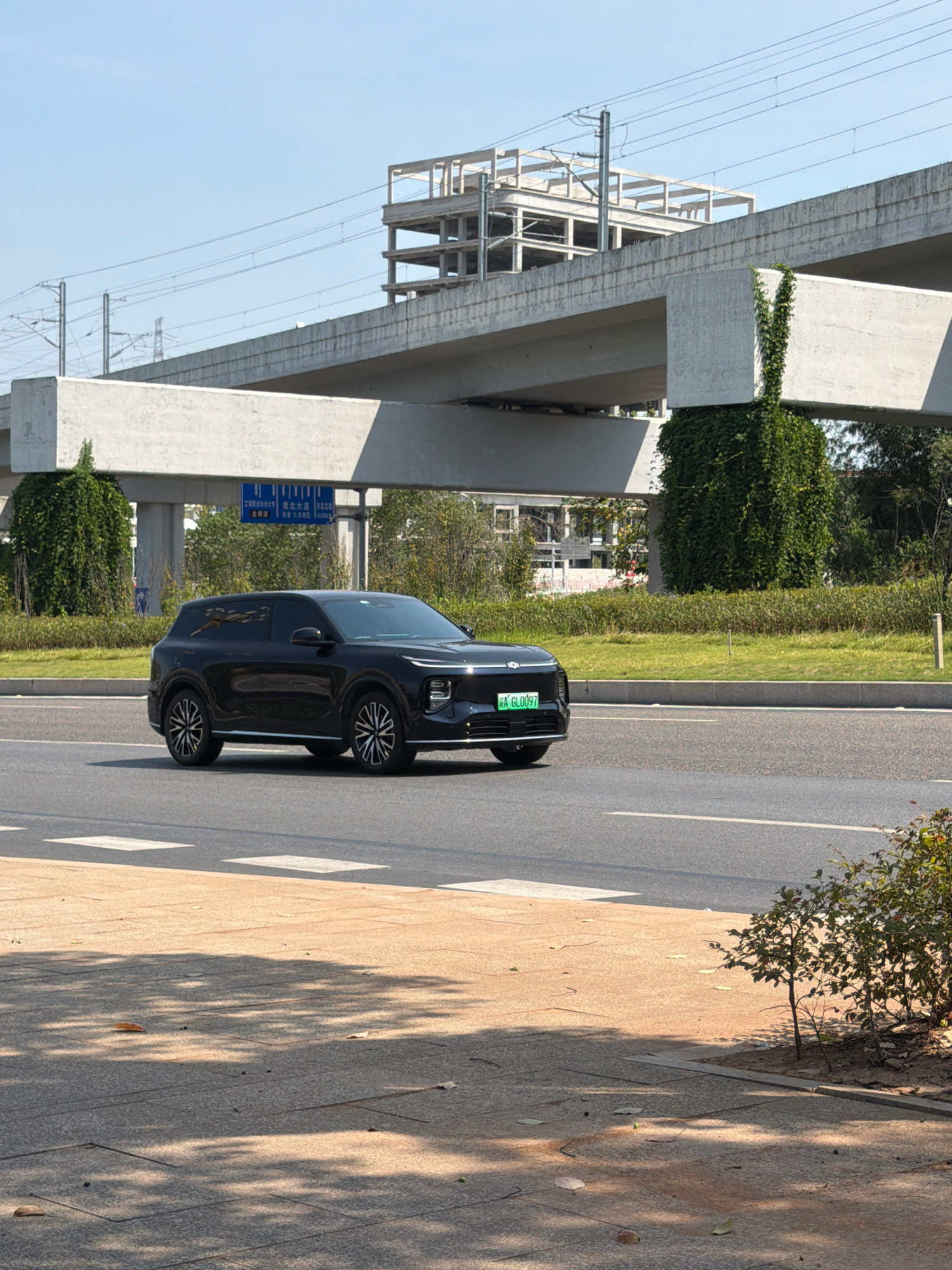 The image depicts a black SUV driving on a city street in Jiangjin, China. The car has a green license plate that reads "A GL0097". In the background, a multi-level overpass, under construction, is visible. The overpass is supported by concrete pillars covered in ivy. The sky is clear and blue. The street is paved with asphalt, and there is a pedestrian walkway with shadows cast by trees. The overall environment suggests an urban setting with ongoing infrastructure development.