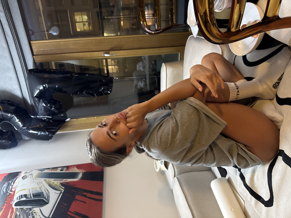 A fair-skinned woman with blonde hair sits on a beige and black patterned couch, her right arm bent, hand touching her face. She wears a gray t-shirt and white socks with stylized lettering. Behind her, a window reflects a room with wooden cabinets. Black balloons shaped like numbers are to the left, and a red and black modern art piece is visible in the lower left corner. A curved gold decorative object rests on the couch's back.