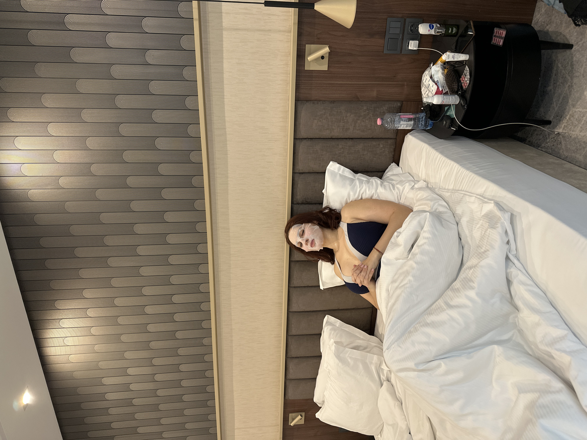 A medium shot of a woman lying in a bed. She is on her back under a white comforter with multiple pillows. She has a white face mask on and is wearing a blue bathing suit top. Her hands are folded on her chest. Next to the bed is a black, round table with many items on it, including a water bottle, a white bottle with a yellow top, a black bag, pills in packaging, a hair brush, and other items. The wall behind the bed is a combination of brown wooden panels, gray upholstered panels, and gray wallpaper with black and silver designs. The lamp is positioned above. The room appears to be a hotel room in Kazanlak, Bulgaria. The specific location within the city cannot be determined from this image.