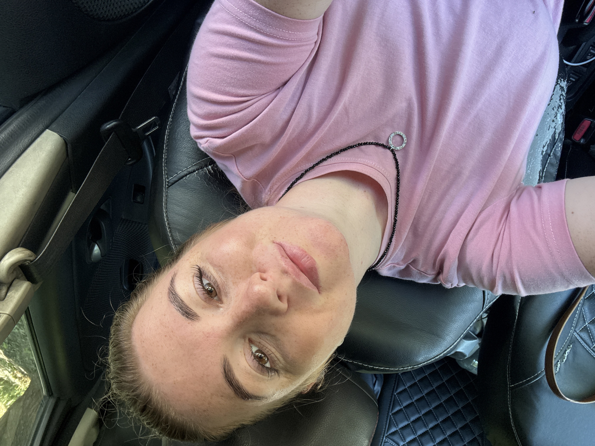 A woman with light skin and freckles is sitting in the passenger seat of a vehicle. She has blonde hair pulled back, and she is wearing a pink t-shirt and a black necklace with a circular pendant. The interior of the car is visible, including black leather seats and a seatbelt. The scene appears to be outdoors, with sunlight filtering in.
