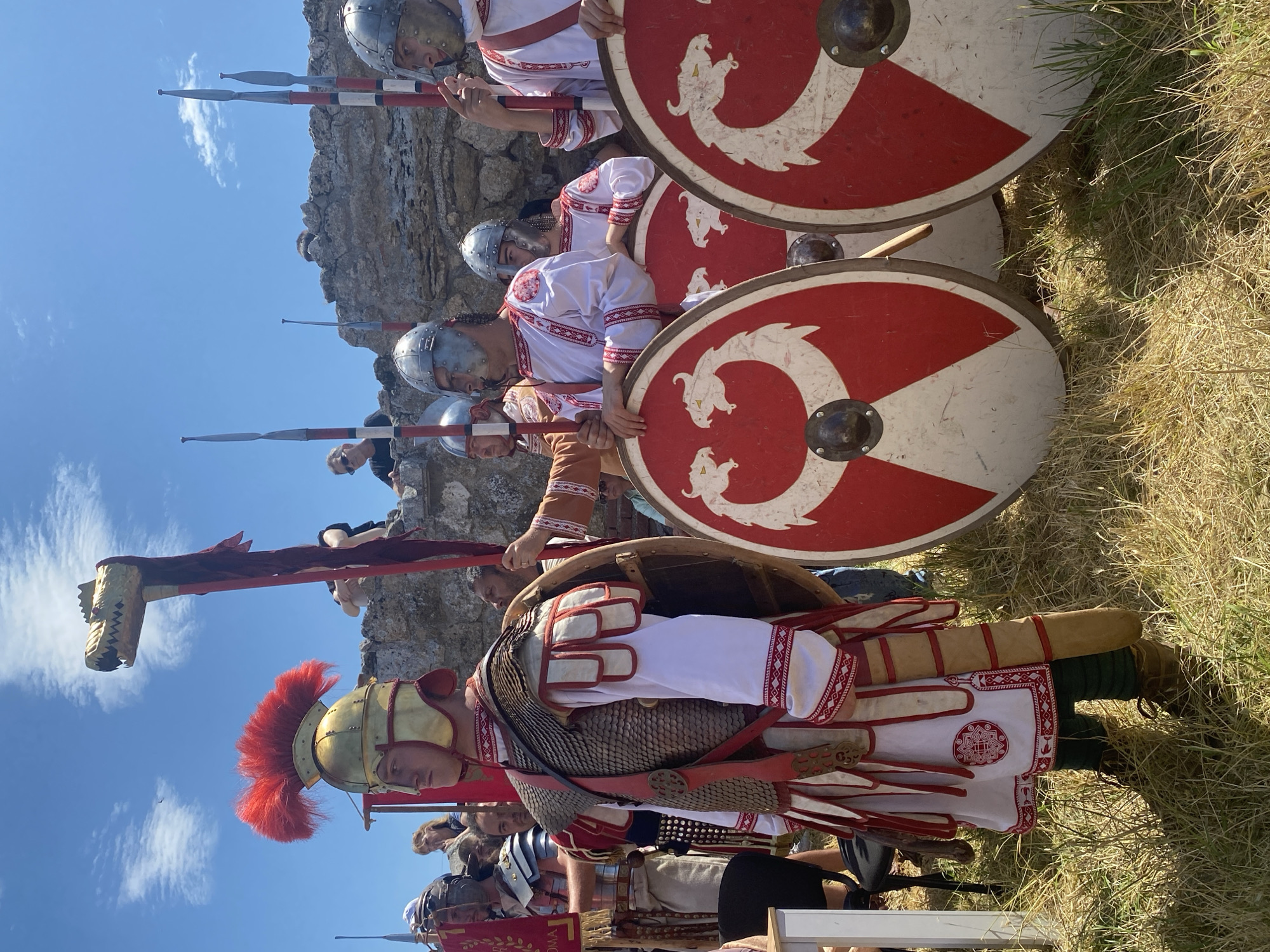 This detailed description pertains to an image captured in Gigen, Bulgaria.
The image depicts an outdoor scene featuring several individuals in historical military attire, standing in what appears to be a formation or display. The sky is bright blue with scattered white clouds, indicating clear, sunny weather. The foreground and midground consist of dry, straw-colored grass, characteristic of a warm, possibly summer, environment.
In the left-center, a man stands prominently, facing left from the viewer's perspective. He wears a gold-colored helmet with a large, vibrant red horsehair crest. His body is covered by a mail shirt (lorica hamata), a white tunic with intricate red geometric embroidery along the cuffs and hem, and white segmented leather pteruges on his shoulders and lower torso. A red belt with ornate metal disc decorations cinches his waist. His lower body is clad in dark green leggings or trousers, and he wears brown lace-up boots. A brown wooden shield is partially visible on his left side.
Behind this central figure and slightly to his right, another person, largely obscured, holds aloft a tall standard topped with a gold-colored, stylized dragon or serpent head (a draco). A flowing red fabric streamer extends downwards from the standard.
To the right of the central figure, a line of at least four other individuals stands, also in military costume. They wear silver-colored helmets and white tunics adorned with the same red geometric embroidery as the main figure. Each holds a spear (hasta) with a dark tip, and red and white striped shafts. They also carry large, round shields. These shields feature a predominantly red field with a central white inverted triangle or 'V' shape. Within the red sections, white stylized dragon or serpent heads, open-mouthed, are depicted facing each other. The shields have a central, dark-colored metal boss and appear somewhat worn.
In the background, behind the costumed individuals, a rough, light-colored stone wall rises, and several civilian spectators are visible observing the scene. Some appear to be looking over the wall, while others stand further back. They are dressed in modern casual clothing. A partial view of a white table and black chair is visible on the far left of the frame.
