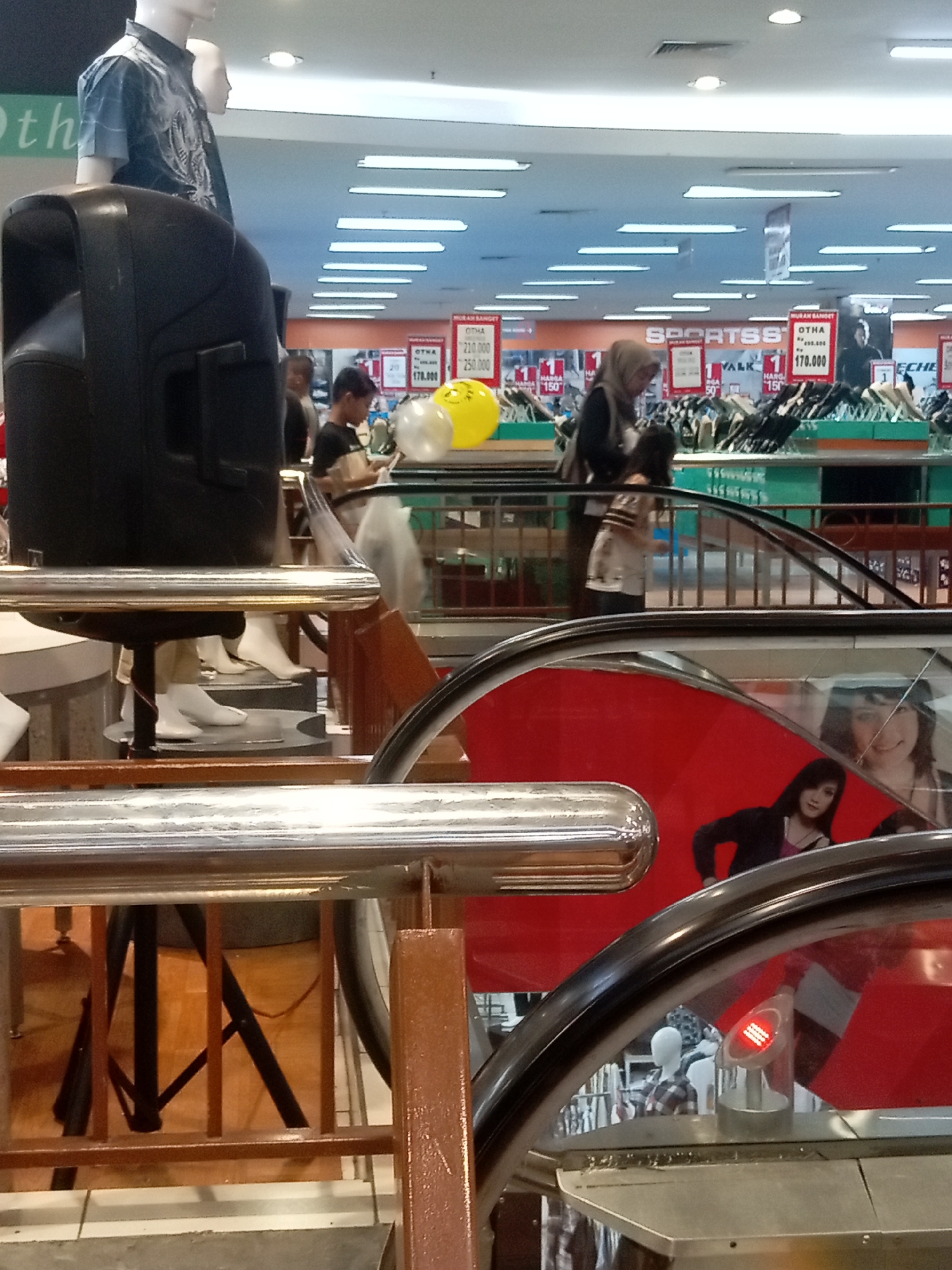 The image depicts the interior of a department store, likely a Matahari department store, in Batam, Indonesia. The foreground features a black speaker on a stand. Behind it, a mannequin is dressed in a short-sleeved, dark patterned shirt. Further back, an escalator is visible with people interacting with it. In the midground, two balloons, one clear and one yellow, are floating near the ceiling. Various store displays and signage are present, including signs for "SPORTS" and promotional pricing like "170.000". In the background, mannequins and shelves of merchandise are visible, illuminated by fluorescent lighting. The overall environment suggests a commercial shopping space.