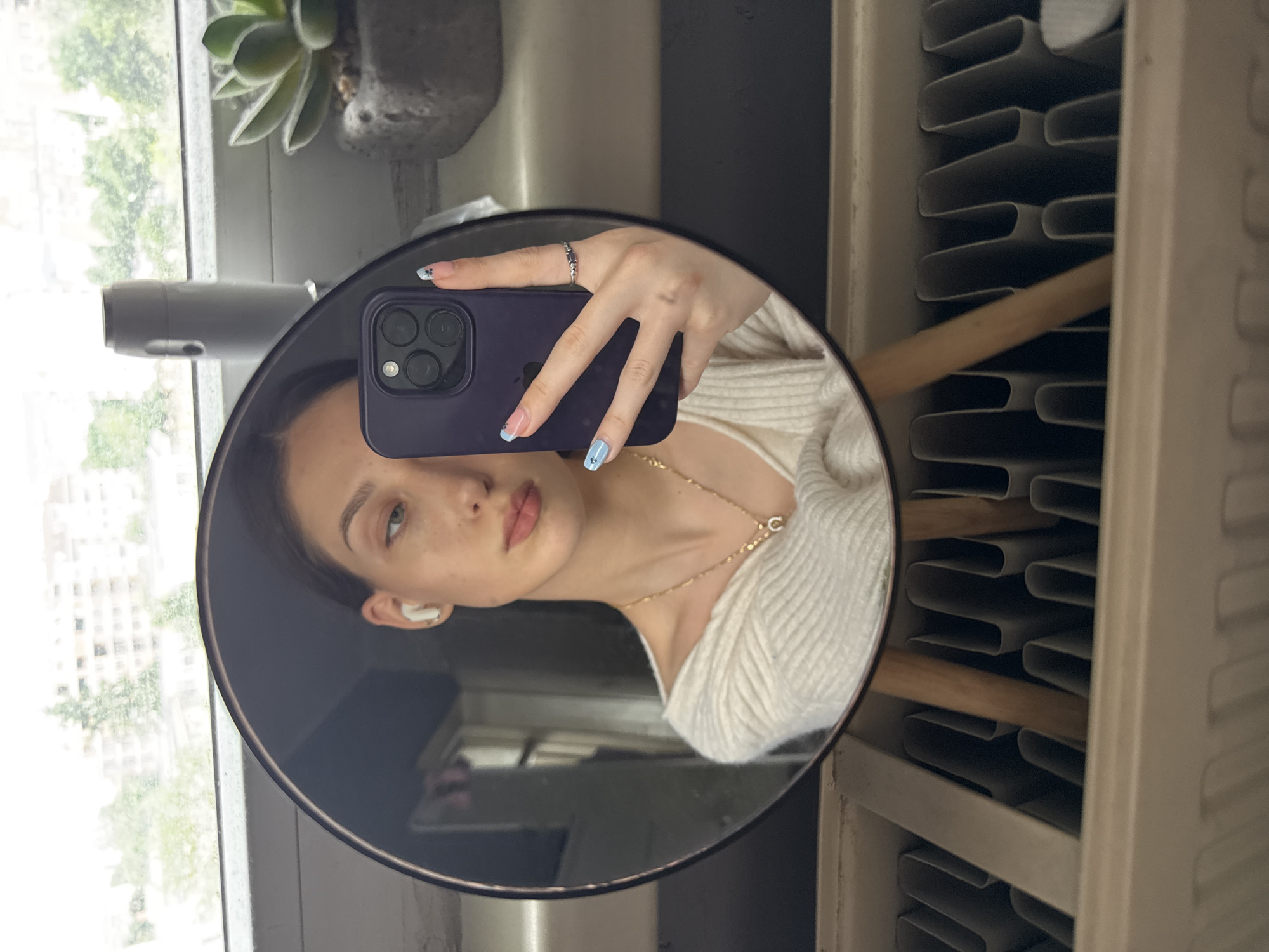 A mirror selfie features a woman with fair skin, light brown hair pulled back, and wearing a white wireless earbud. She holds a purple iPhone 14 Pro to take the picture, displaying manicured nails with a blue and white design and a simple ring on her finger. The reflection reveals a white, textured sweater and a delicate gold necklace with a small pendant. The background behind the mirror includes a window with a view of buildings and greenery, a succulent plant in a gray pot, a gray cylindrical object, and a white radiator with wooden dowels. The scene suggests an urban setting, with the window possibly overlooking a street in Sofia, Bulgaria. The overall lighting is soft and natural.