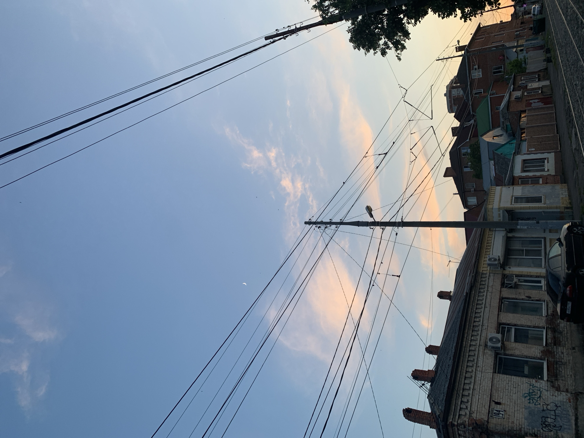 The image depicts a street scene at dusk in a city. The sky is a gradient of pale blue to soft orange and pink hues, with wispy clouds and a visible crescent moon. A dense network of overhead wires, likely for electrical transport like trams or trolleys, crisscrosses the frame. Several utility poles support these wires.
In the foreground, a row of old brick buildings lines the street. The building on the left is weathered, with visible graffiti on its facade and a house number "194." It has multiple windows, some with air conditioning units installed, and distinctive brick chimneys on its roof. To the right of this building, there is a dark-colored sedan parked.
Further down the street, more buildings of varying sizes and architectural styles are visible, some with red brick, others with green roofs. The street itself appears to be paved, with what looks like tram tracks running through it. A large, dark green tree is prominent on the far right side of the image. The overall impression is of an aging urban environment at the end of the day.