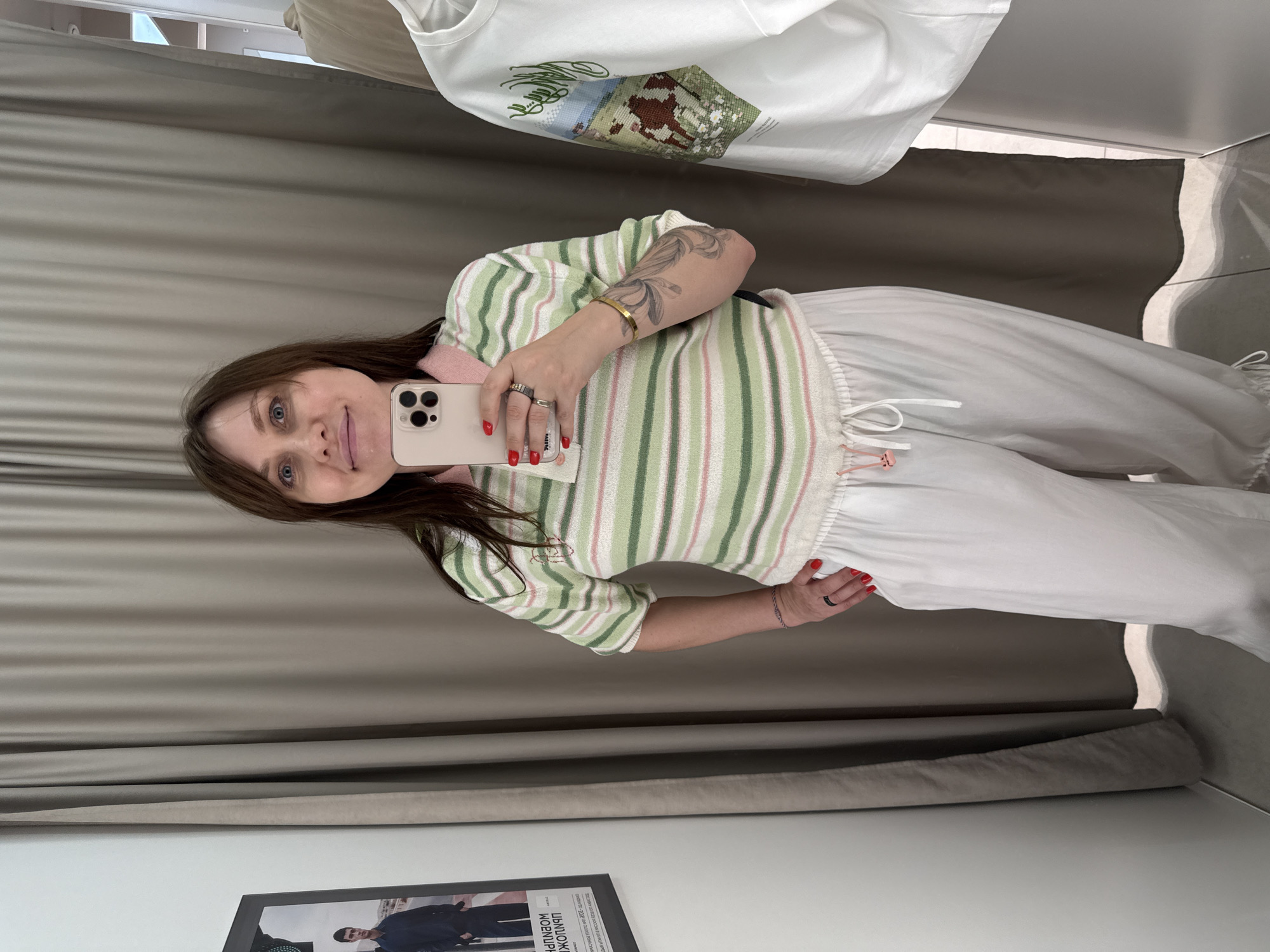 A woman with brown hair and blue eyes is taking a mirror selfie in a fitting room. She is wearing a striped green, white, and pink polo shirt and white linen pants with a drawstring waist. She is also wearing rings on her fingers and a bracelet on her wrist. In the background, there are beige curtains, a poster on the wall, and a white t-shirt hanging on a hanger. The lighting is bright, and the overall atmosphere appears to be in a retail store. The poster on the wall has Cyrillic text, and it is possible this fitting room is located in Sochi, Russia, given the context provided.