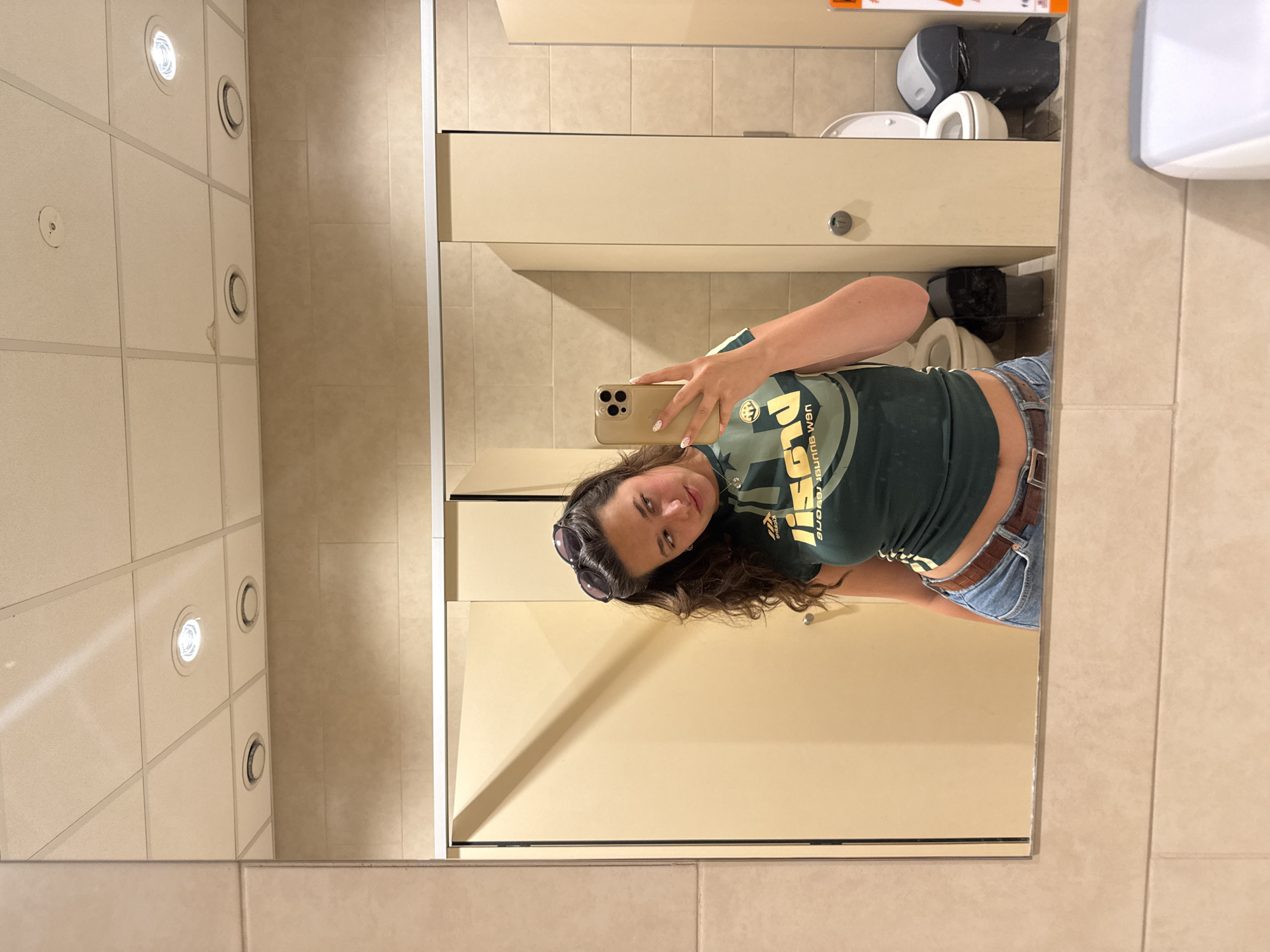 A woman is taking a selfie in a bathroom mirror. She is wearing a green t-shirt and jeans with a brown belt. The bathroom has tiled walls and a tiled ceiling with recessed lighting. There is a toilet and a sink in the background. The location is most likely not in Hadzhidimitrovo, Bulgaria, as there is no clear indication of the location.